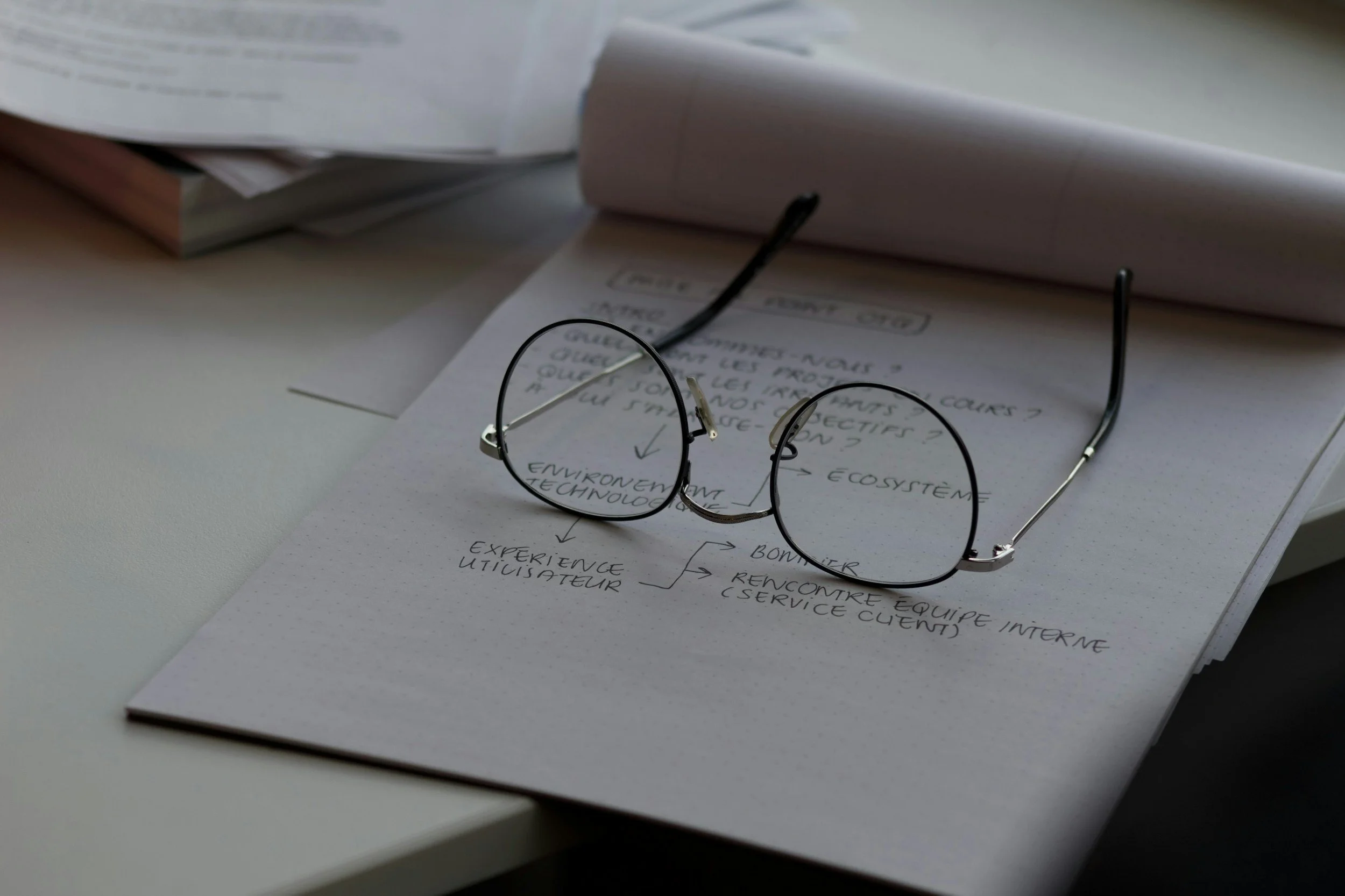 A pair of black-rimmed eyeglasses resting on handwritten notes and documents on a white desk.