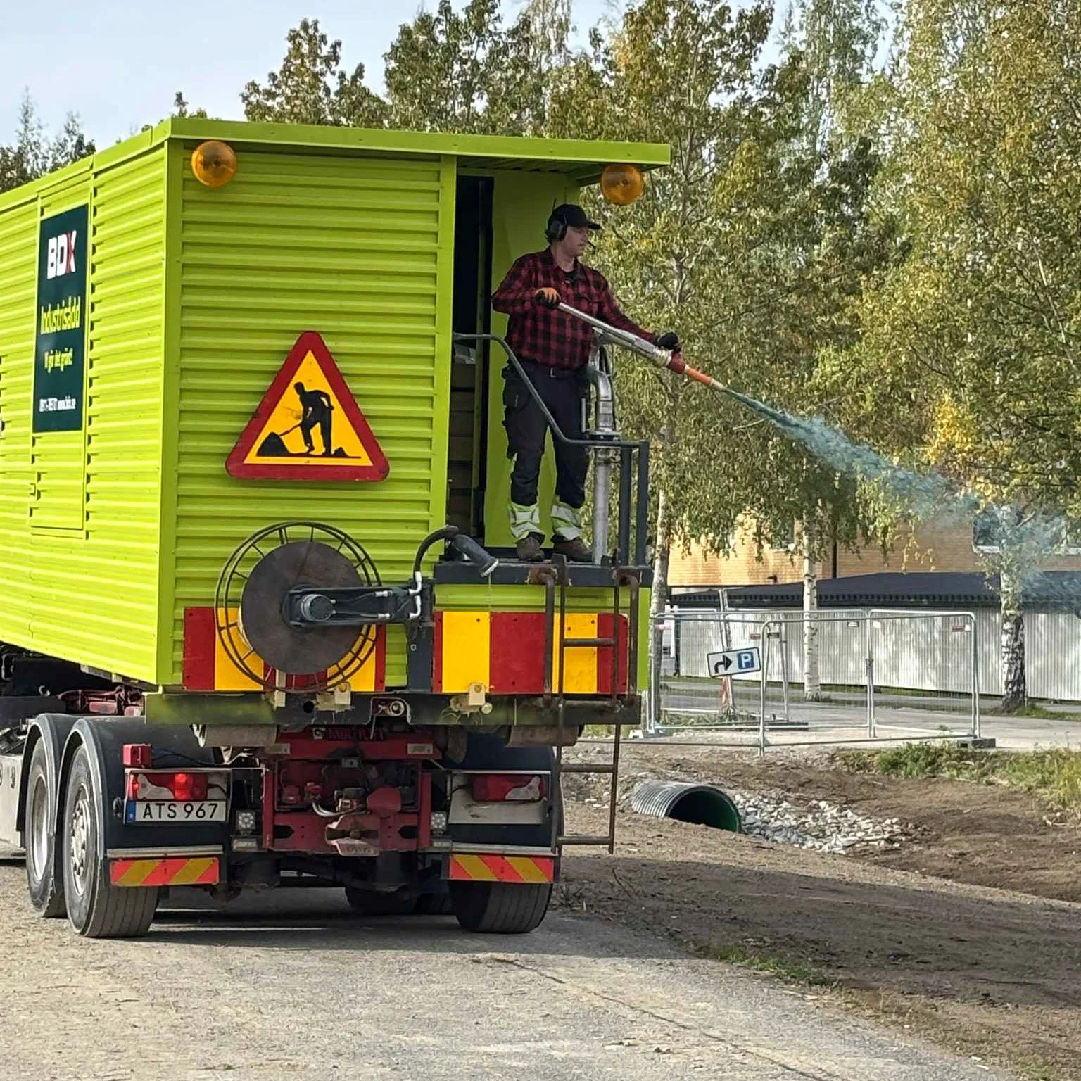 En person i fältarbete som sprutar vatten från ett verktyg, står på en gul och röd transportör med varningsskylt för arbete