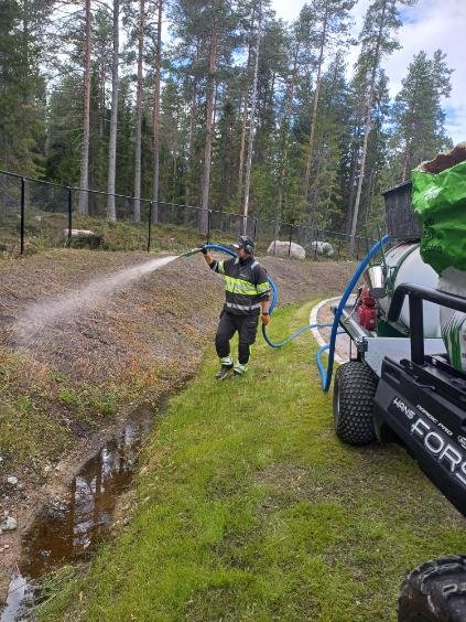 Person wearing a reflective vest and uniform holding a hose, spraying water on the ground in a wooded area with trees and a fence.