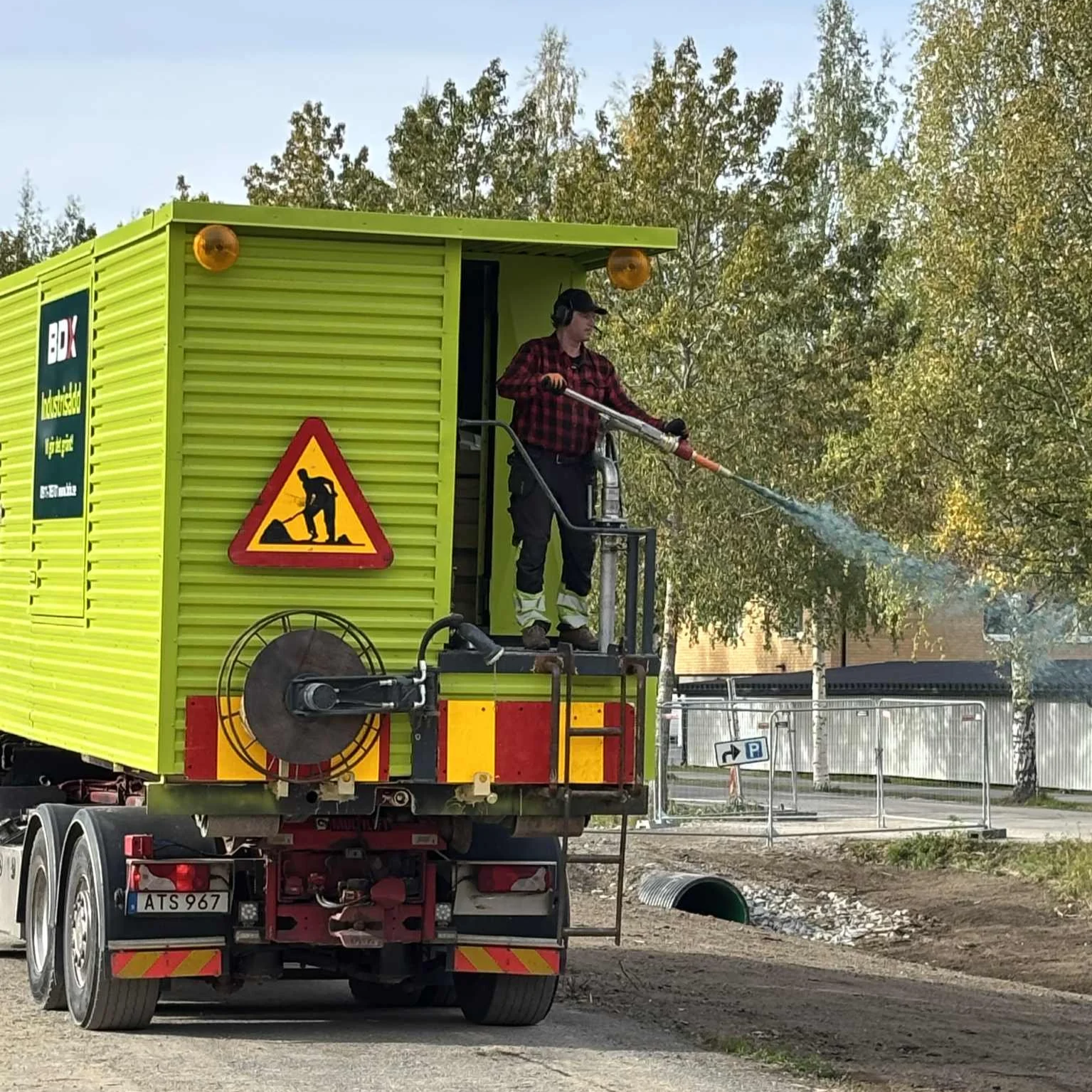 En man står på en höjdarbädd utanför en grön arbetsbil och använder en högtryckstvätt för att rengöra ett rör. Bilen har en varningsskylt med en man som gräver.