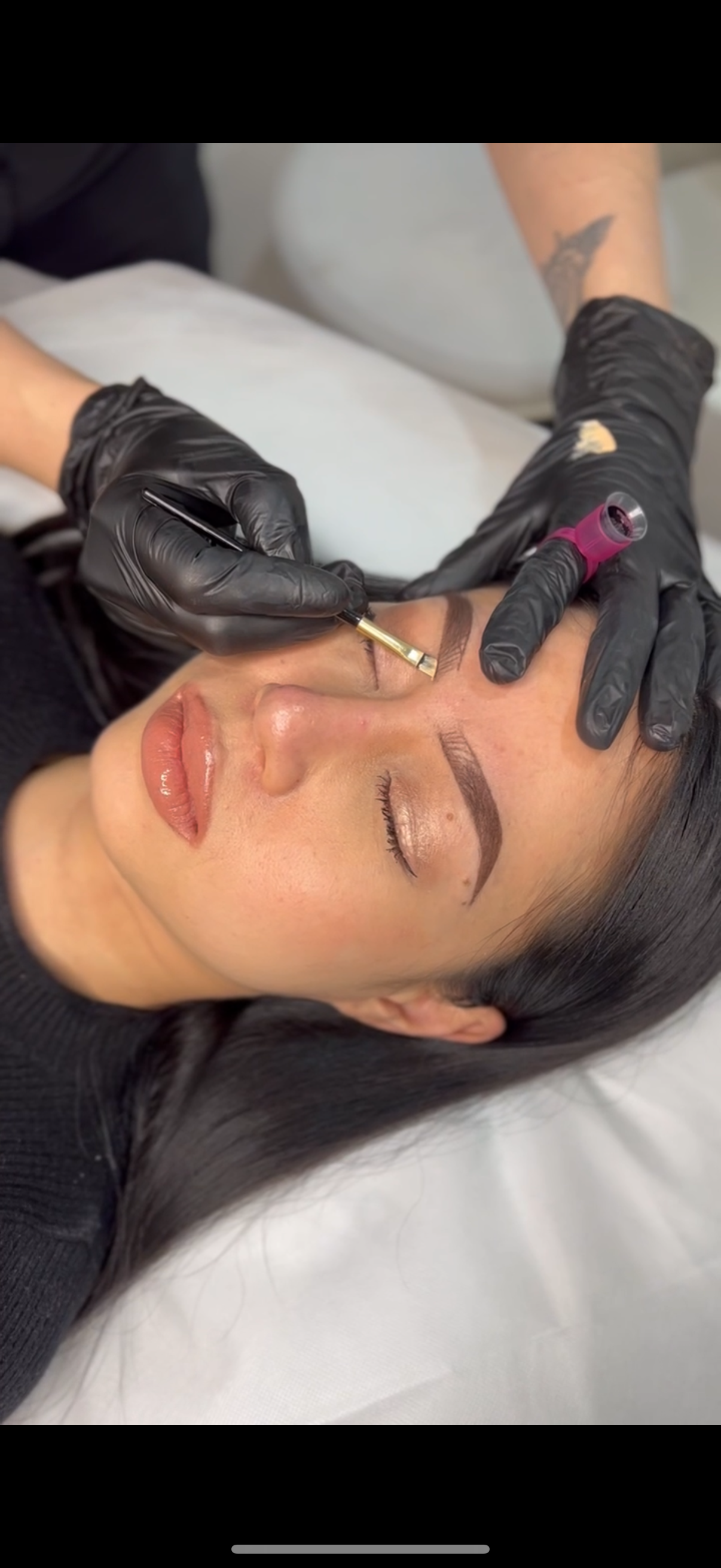 A woman is lying down while a technician is doing a microblading procedure on her eyebrows. The woman has closed eyes, dark hair, and is wearing neutral makeup.