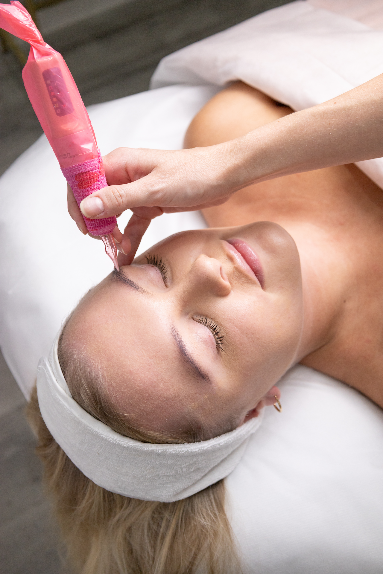 A woman with blonde hair lying on a treatment bed with her eyes closed, receiving a facial treatment from a esthetician using a pink device on her forehead.