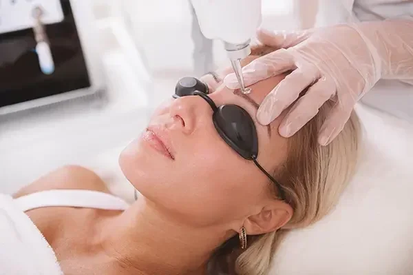 A woman wearing protective glasses receives a facial treatment from a technician who uses a handheld device on her forehead, with gloved hands visible.