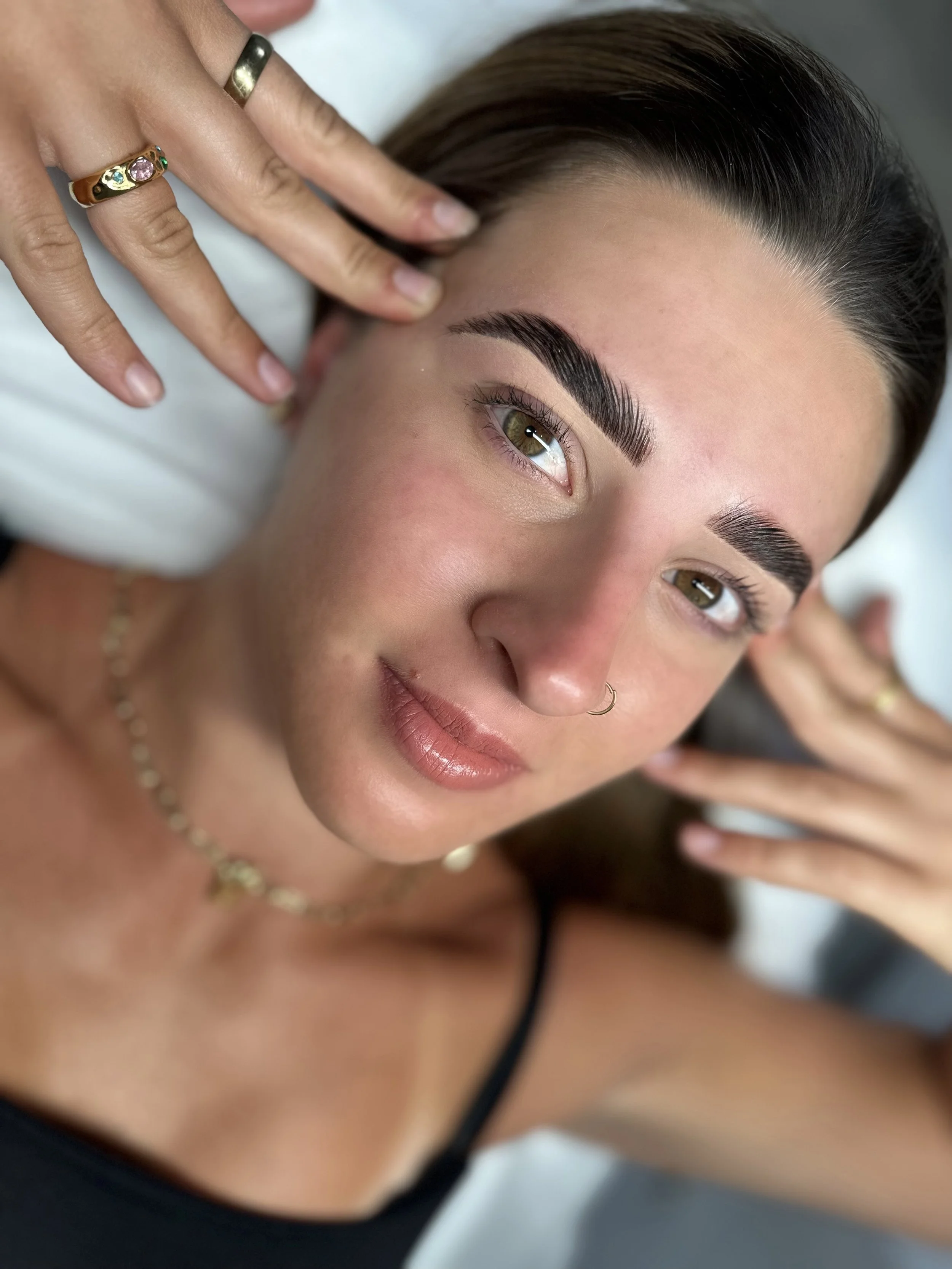 Close-up of a woman with well-groomed eyebrows, wearing a nose ring, necklace, and rings, lying down with her hands framing her face.