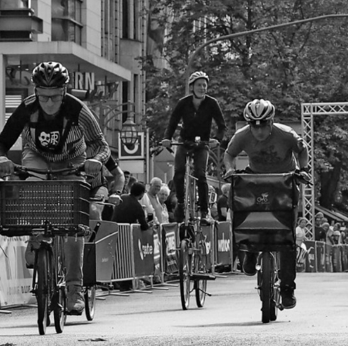 Drei Radfahrer, zwei auf vorderem Platt and einem auf einem Hochrad, fahren in einer städtischen Straße bei einem Veranstaltung mit Zuschauern und Barrieren im Hintergrund.