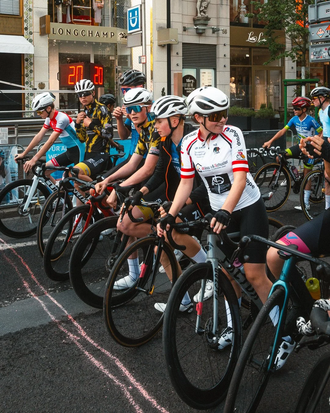 Radsportler in Startaufstellung bei einem Rennen in einer Stadt, mit Gebäuden und Geschäften im Hintergrund.