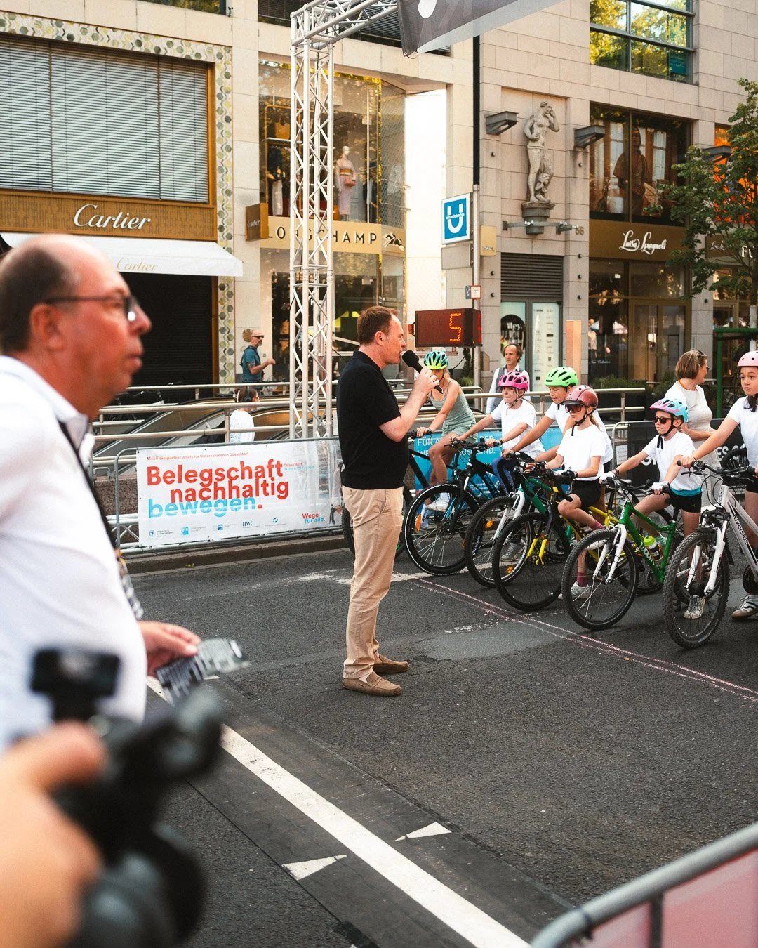 Mann mit Mikrofon spricht zu einer Gruppe von Radfahrern, darunter Kinder mit Helmen, während einer das Rennen anmoderiert. Im Hintergrund sind Geschäfte und eine Uhr auf der Straße sichtbar.