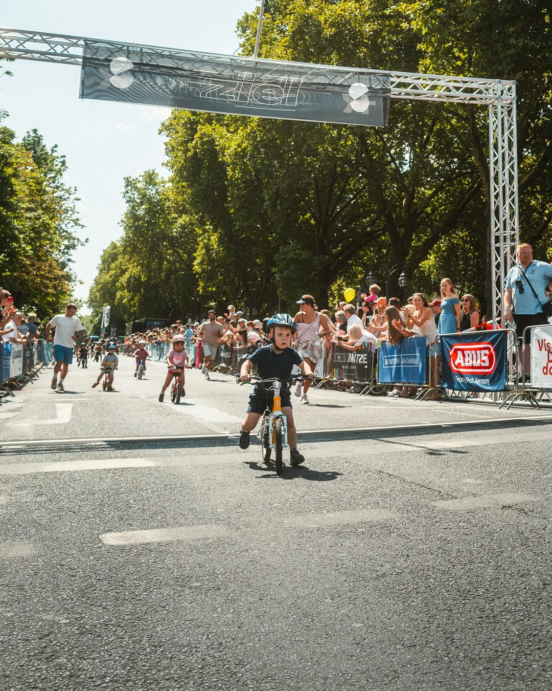 Kinder beim Fahrradrennen auf einer Straße, umgeben von Zuschauern und Bäumen an einem sonnigen Tag.