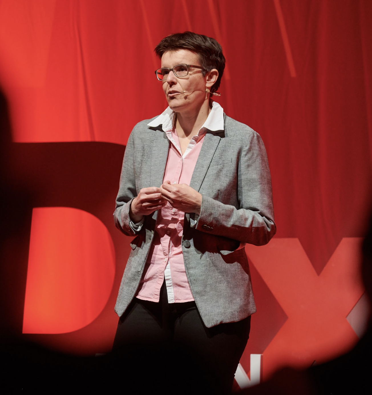 A woman with short dark hair wearing glasses, a gray blazer, and a pink shirt, speaking on stage at a TEDx event with a red background.
