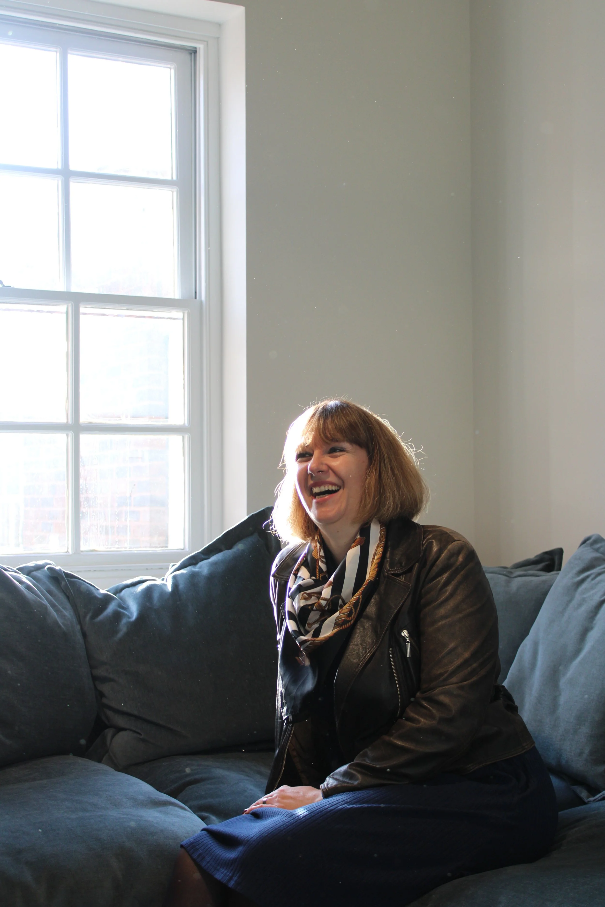 A woman with short brown hair laughing while sitting on a dark blue couch in a room with a large window and white walls.