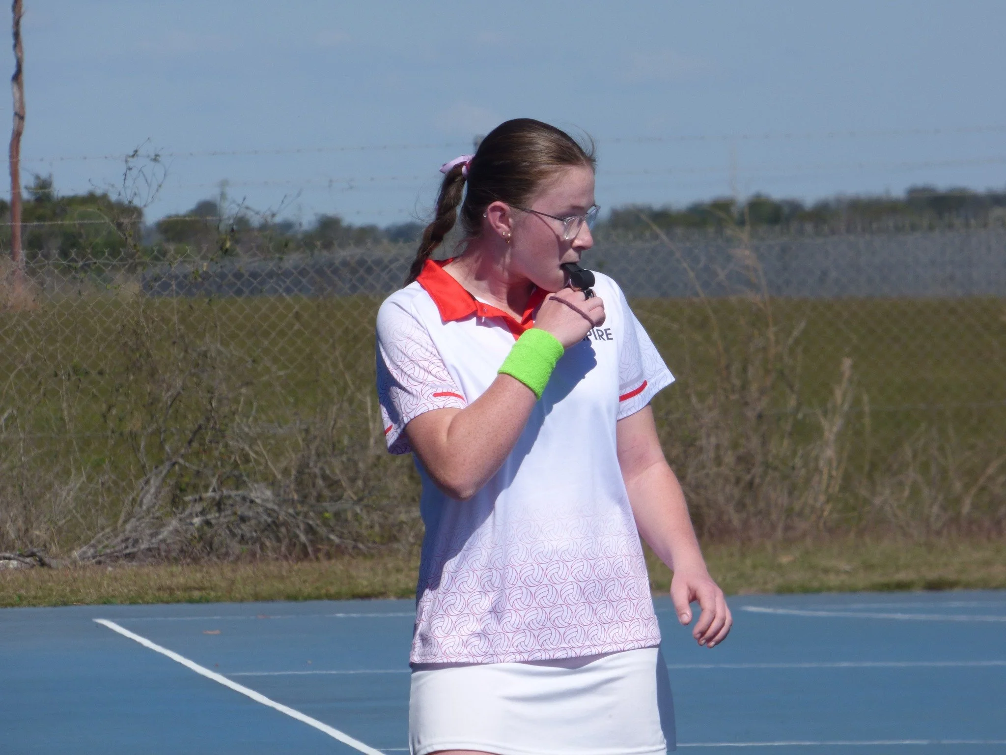 A female netball umpire with glasses, wearing a white and red sports outfit and a neon green wristband, holding a whistle near her mouth on an outdoor netball court.