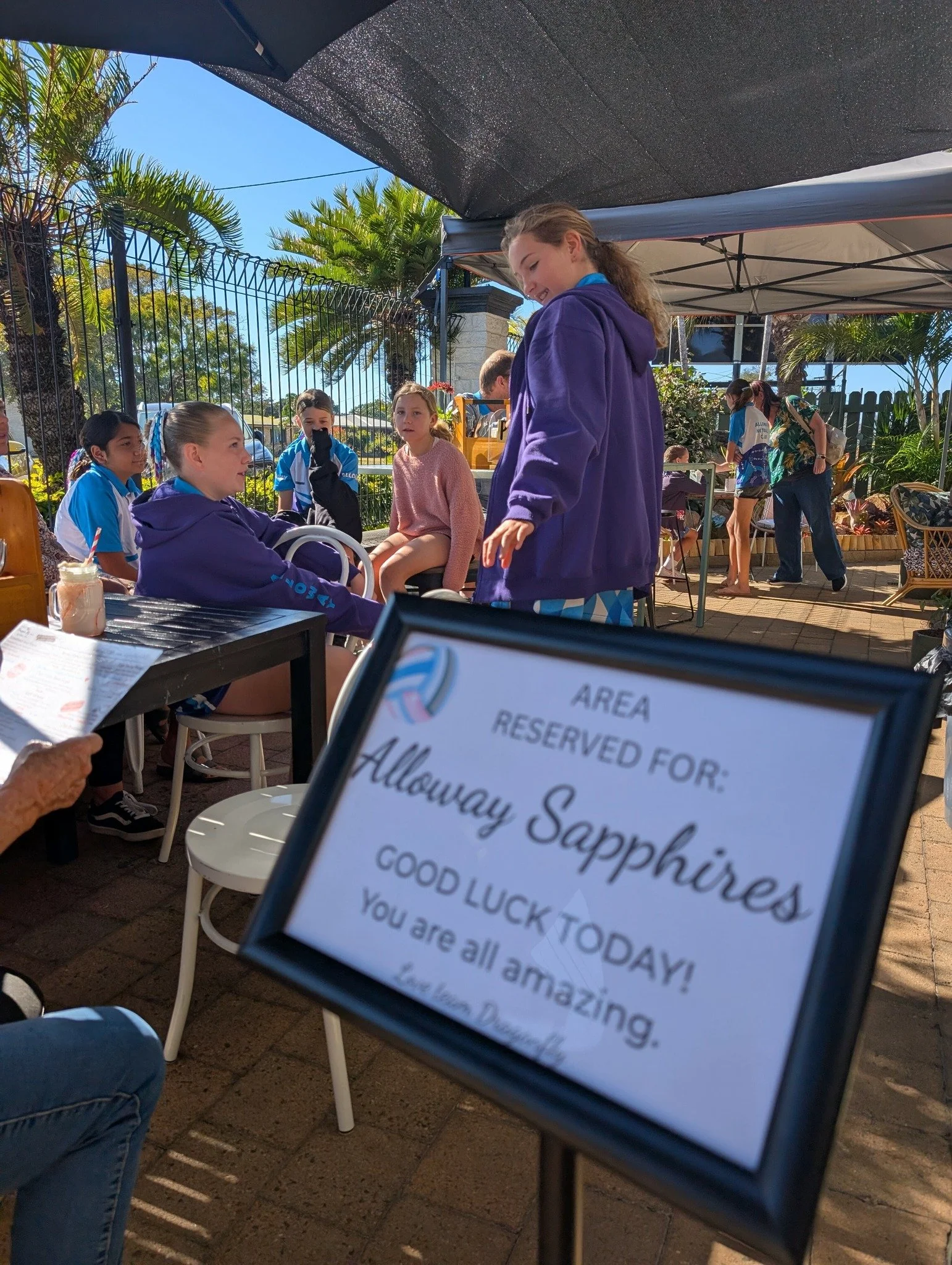 A group of young girls, some wearing purple jackets, sitting and standing outdoors under a shaded canopy, with palm trees and a metal fence in the background. There is a framed sign in the foreground that reads, 'Area Reserved For: Alloway Sapphires. Good luck today! You are all amazing.' The scene appears to be at a team or group event on a sunny day.