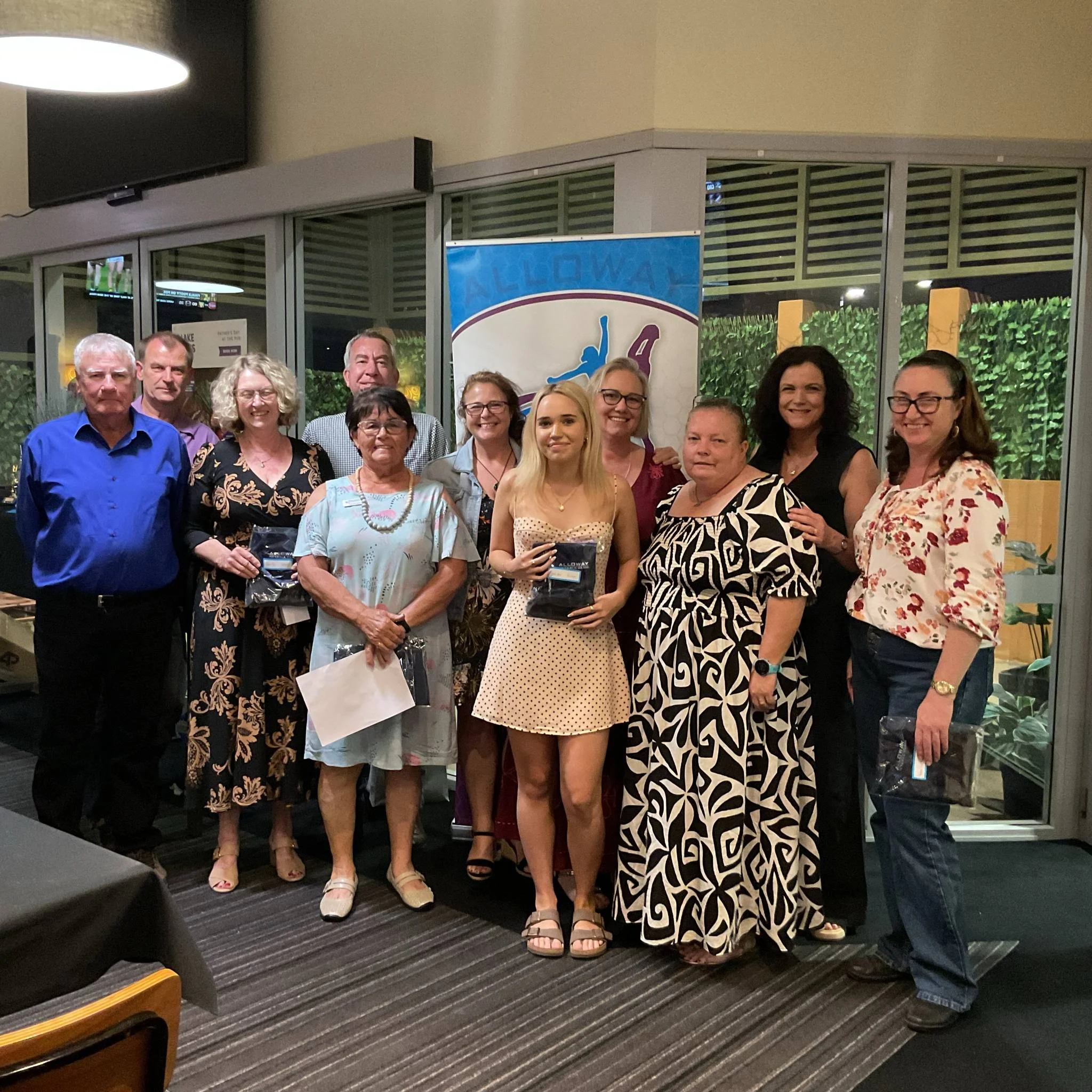 Group of people posing for a photo at a celebration or event, standing indoors near a window with green plants outside, with a blue event banner in the background.