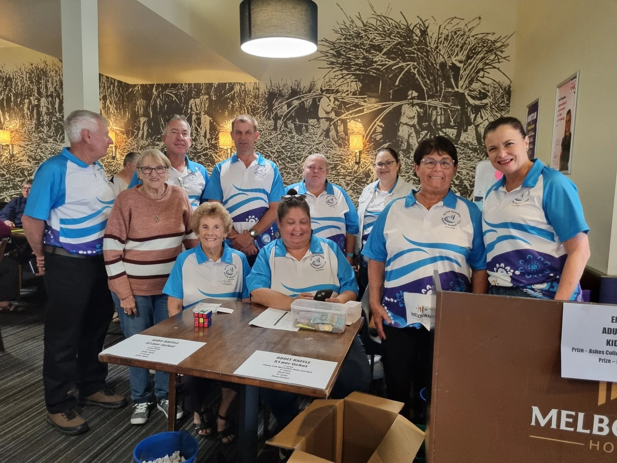 Group of people, some wearing matching blue and white shirts, gathered around a table with raffle signs, in a room with a mural of farm workers harvesting crops on the wall.