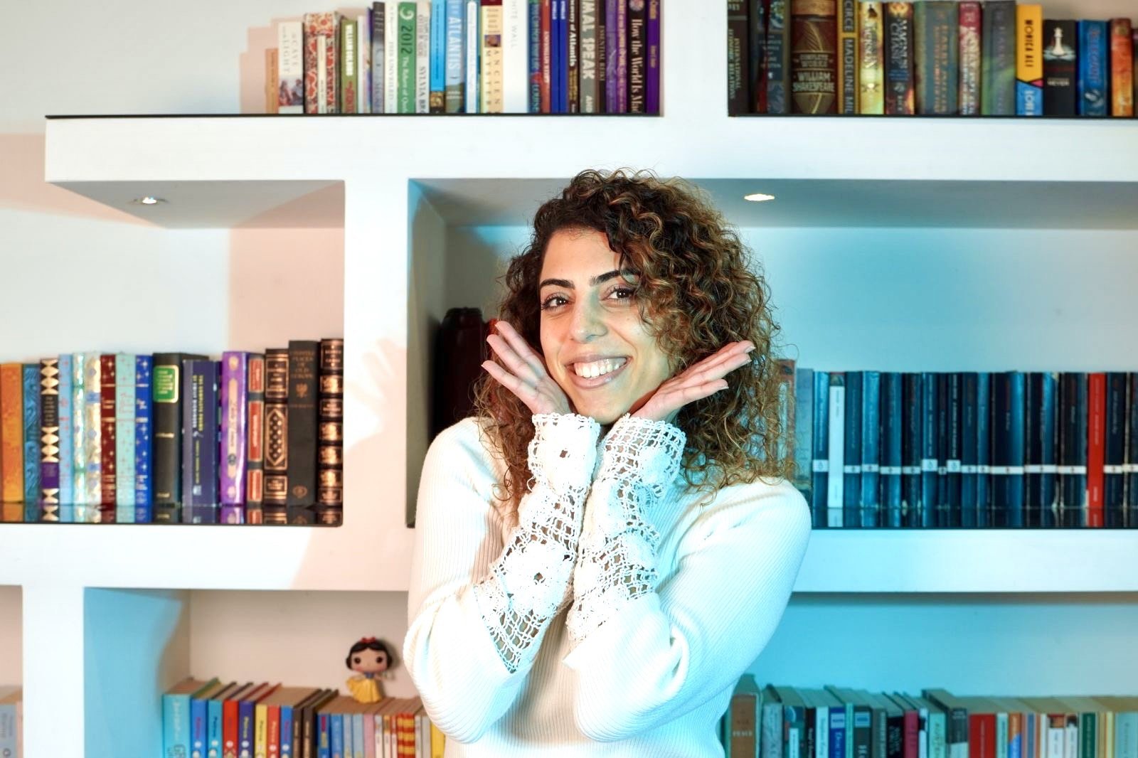 A woman with curly brown hair smiling and posing with her hands under her chin in front of a white bookshelf filled with colorful books.