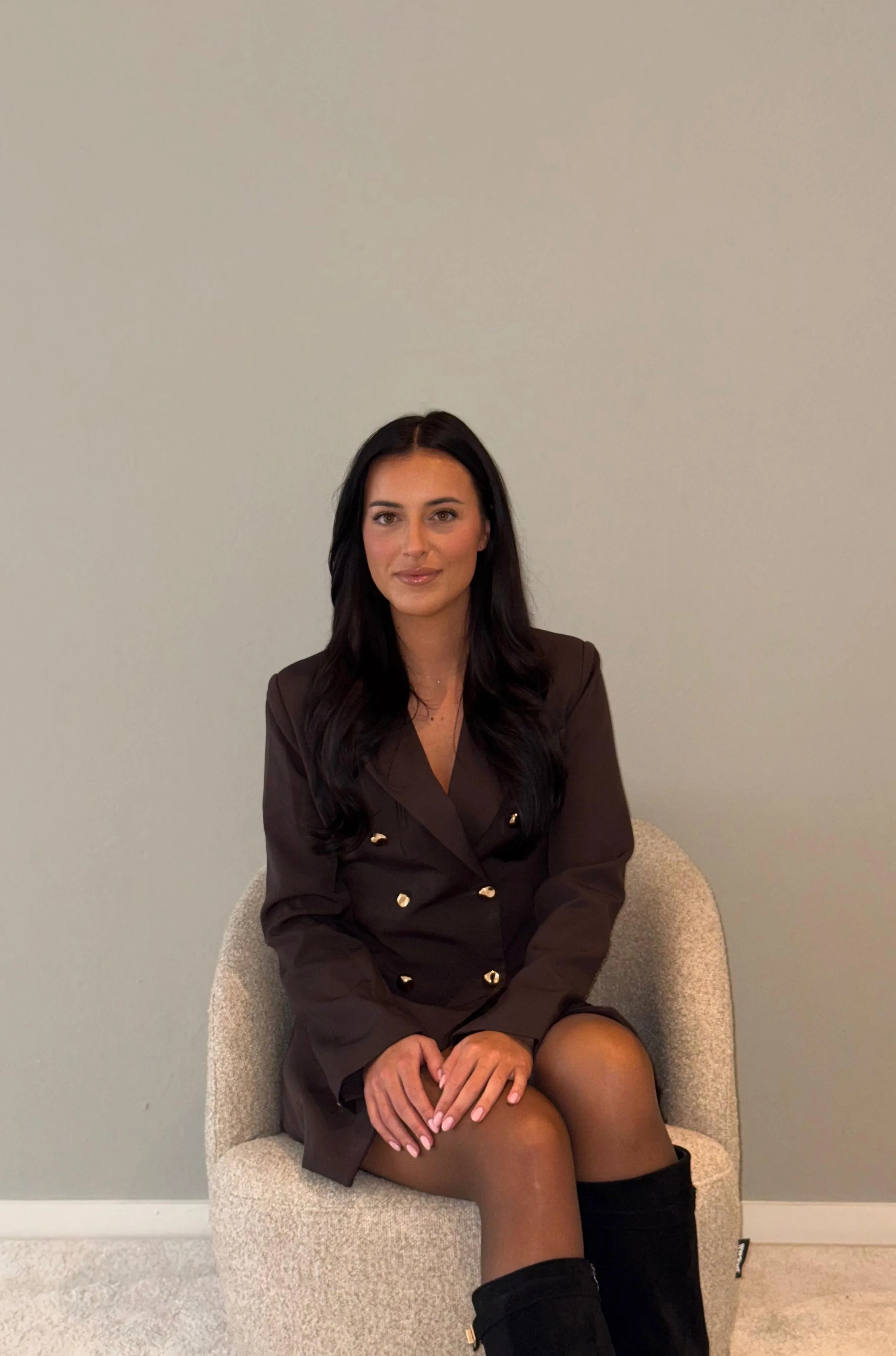 Een jonge vrouw met lang, zwart haar zit op een beige stoel voor een effen lichtgrijze muur, gekleed in een donkerbruin pak en zwarte laarzen.