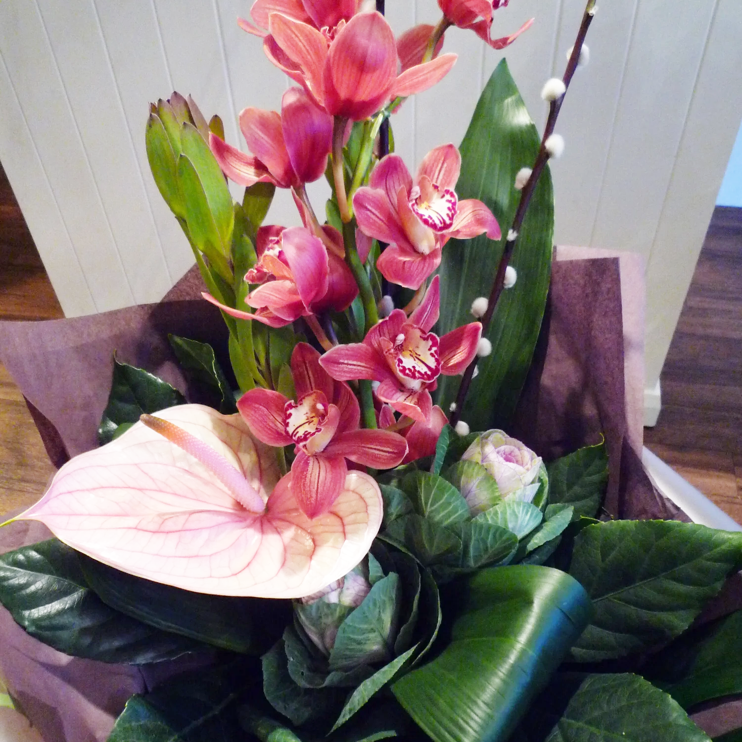 Arrangement of tropical flowers including pink orchids, an anthurium, and other green foliage in a decorative wrapping.