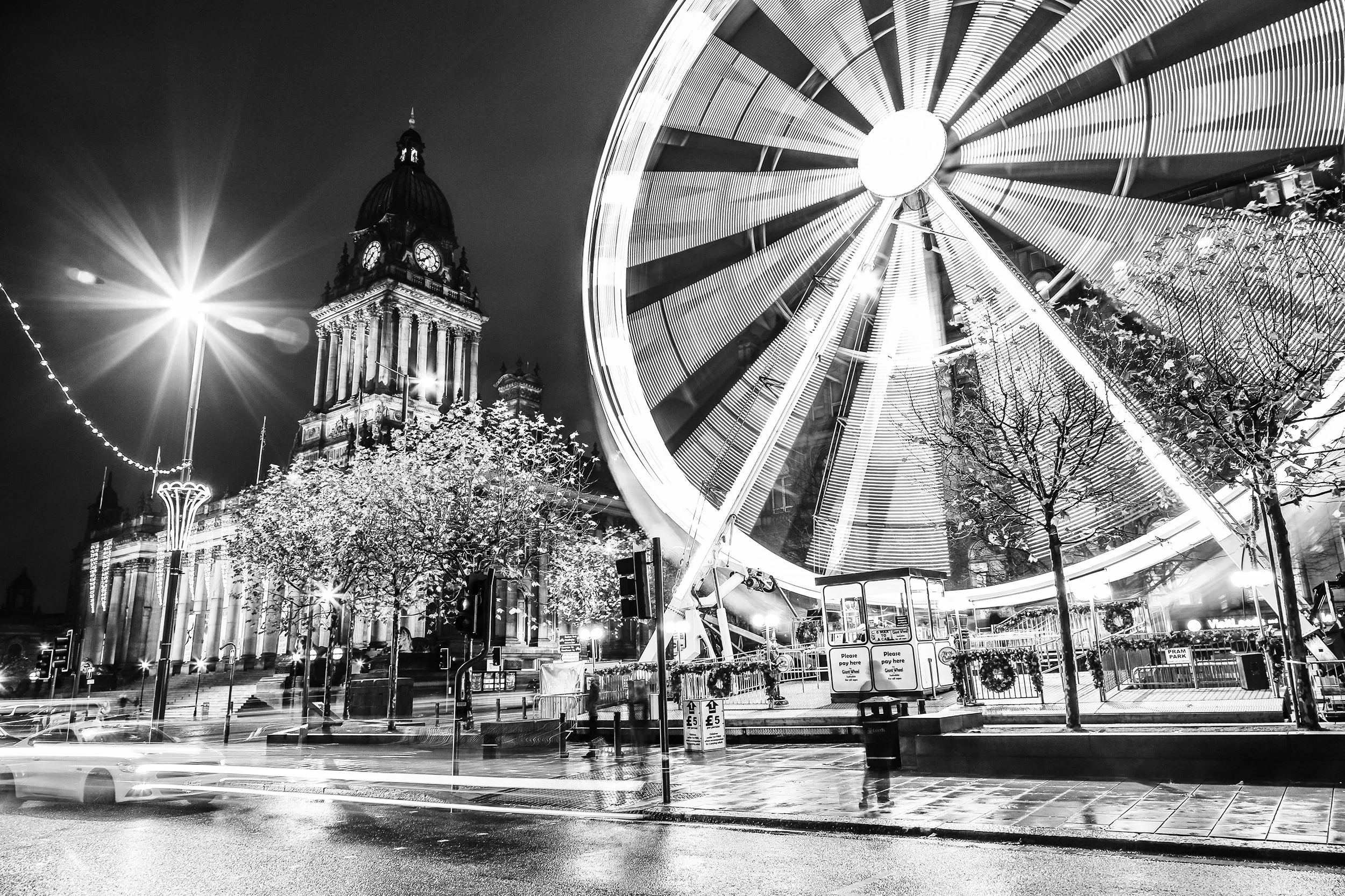 Night scene of a city square with a historic clock tower and a large illuminated Ferris wheel, with trees and streetlights in the foreground, and light trails of passing cars.