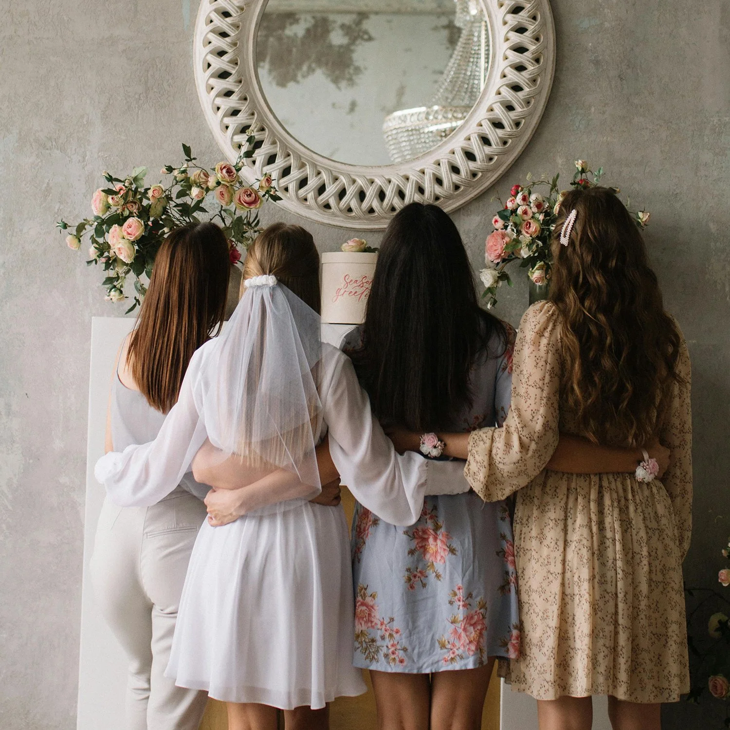 Four women standing together, facing a table with a small white box that has pink writing, in a room with large floral arrangements and a round mirror on the wall.
