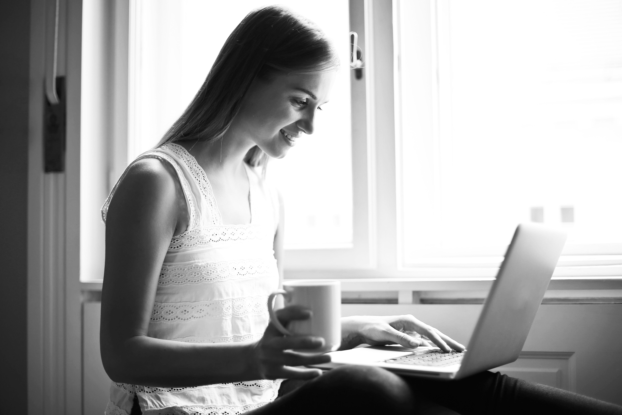 A young woman is sitting at a desk near a window, smiling as she looks at her laptop, holding a coffee mug in her right hand.