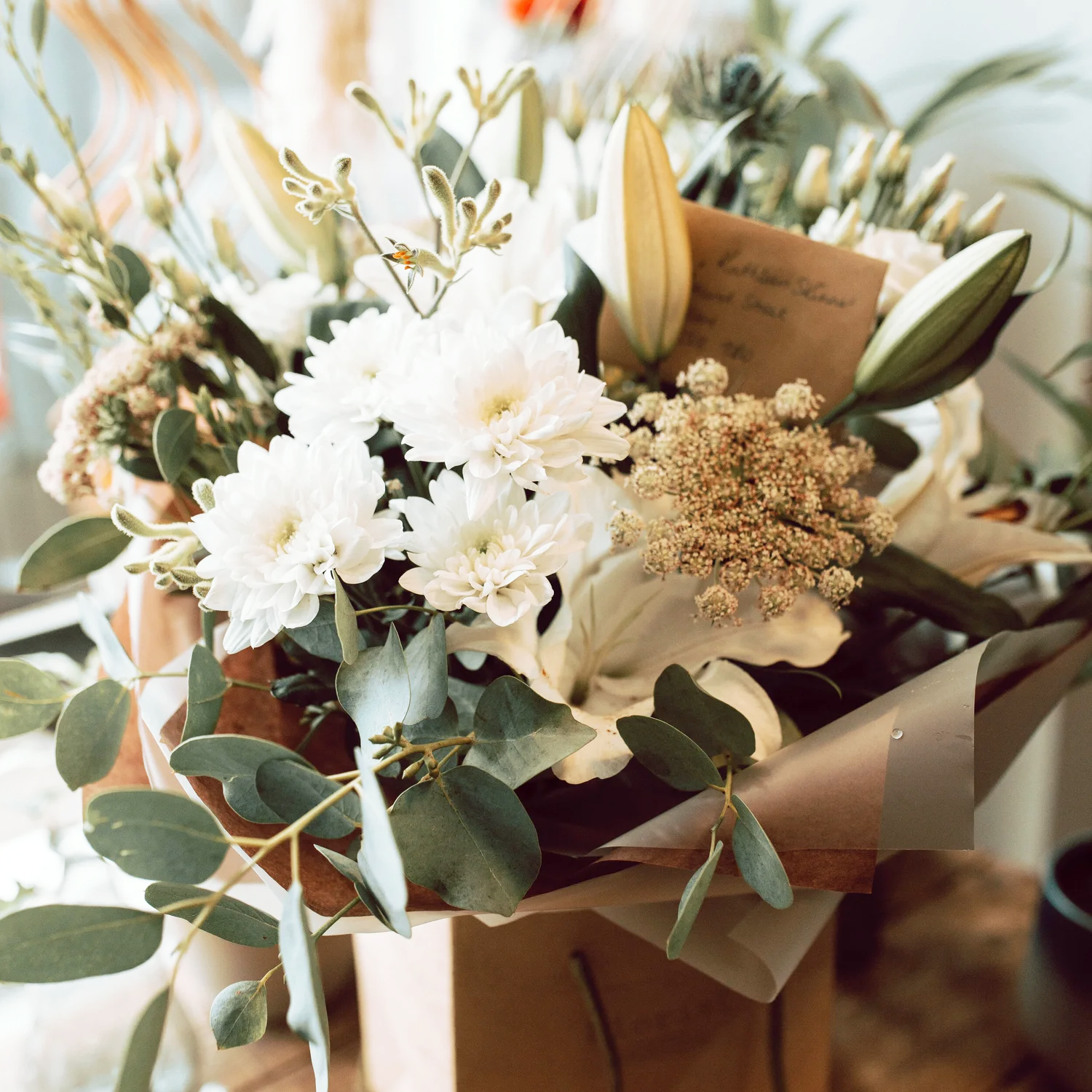 A floral bouquet with white flowers, green leaves, and a handwritten note among the flowers.