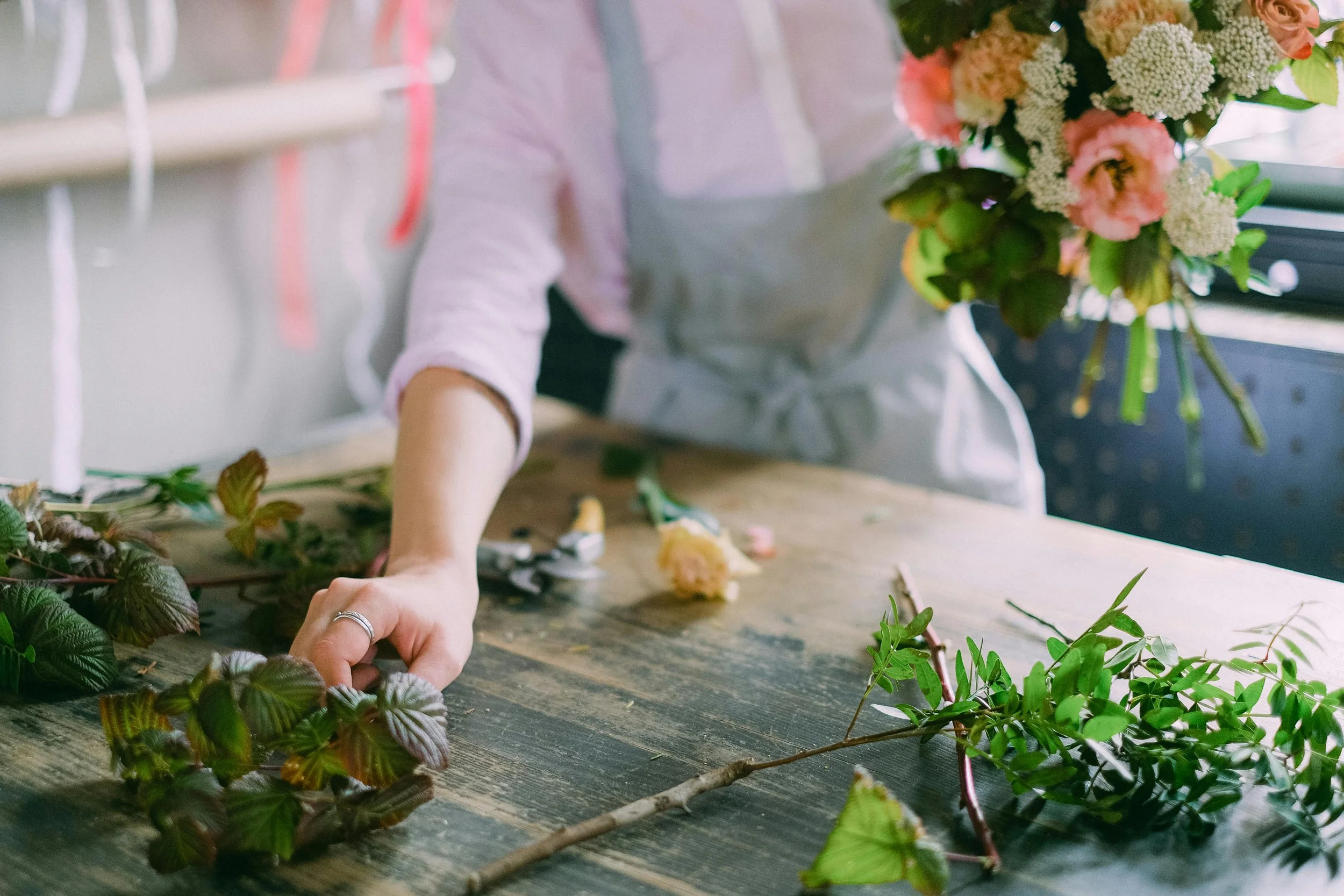 Person arranging a bouquet of flowers on a table with various greenery and floral stems.