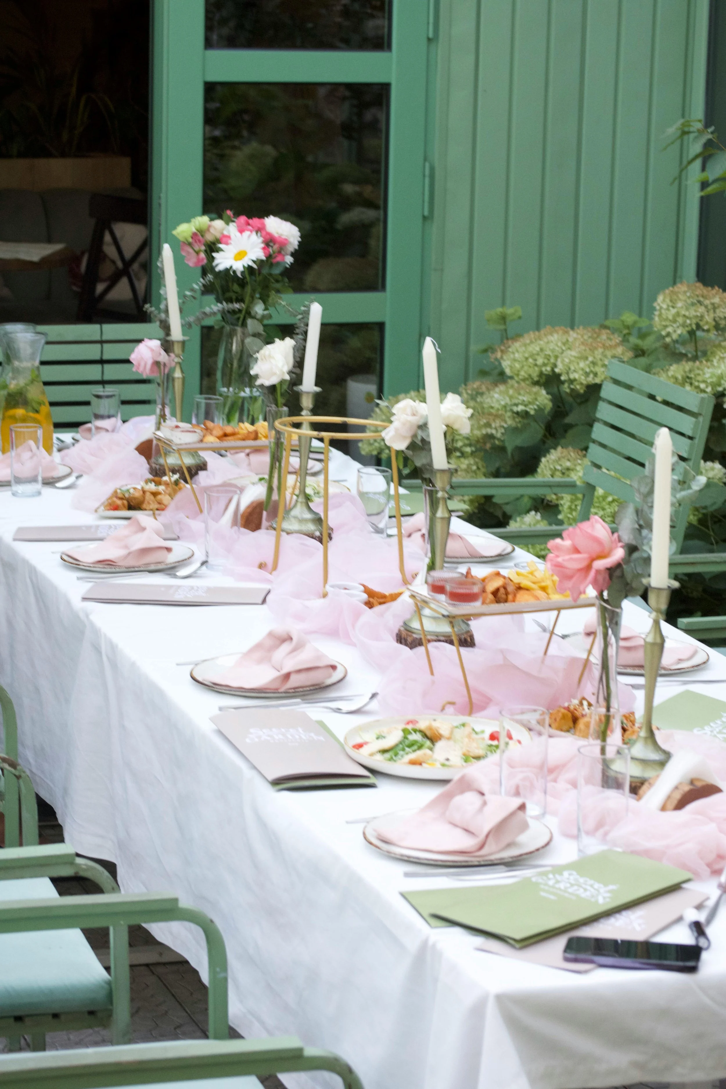 A dining table set for a meal outdoors with pink and white flowers, white candles, plates, glasses, and food, with green chairs and a green door in the background.
