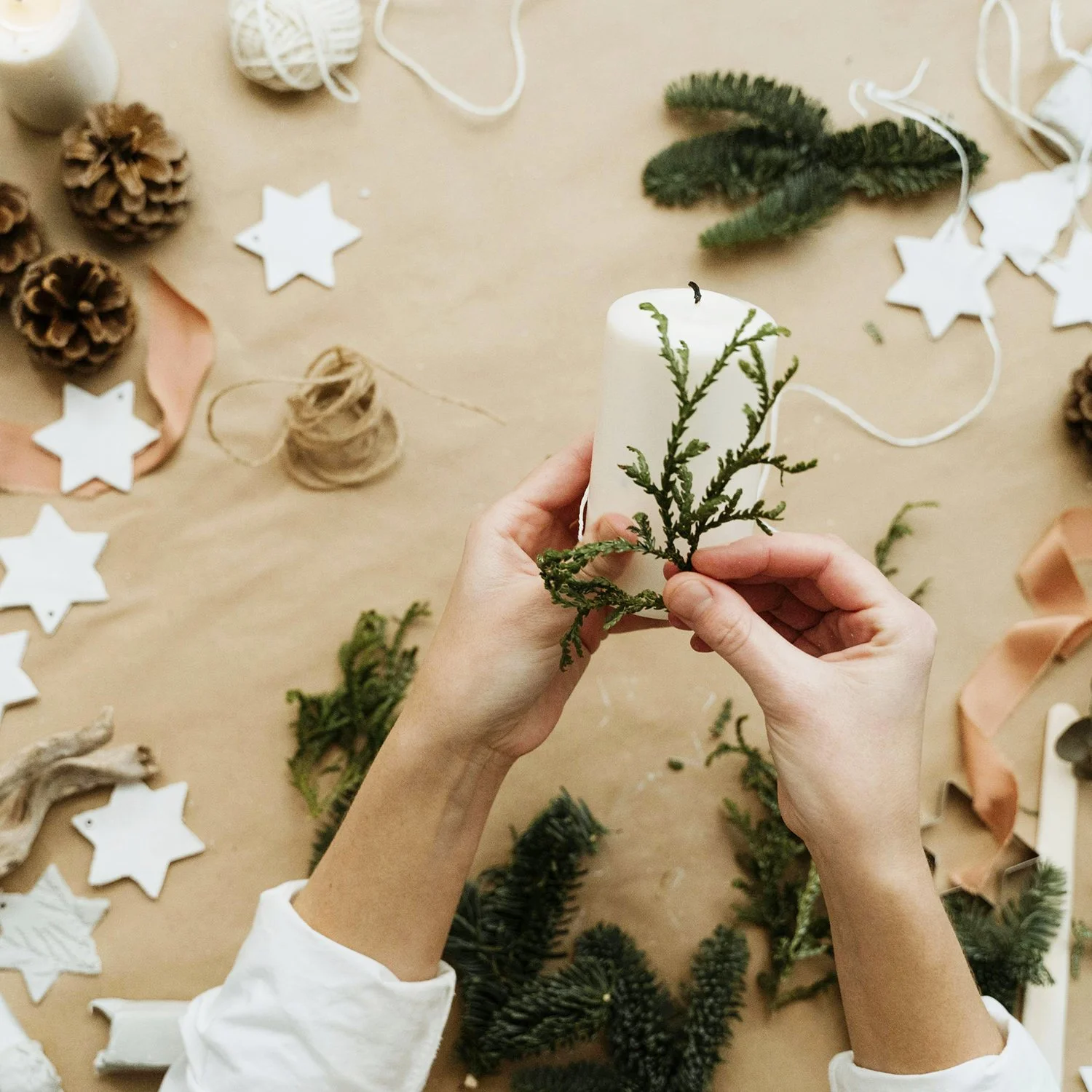 Person holding a white candle decorated with green pine branches surrounded by pinecones, star-shaped ornaments, and crafting materials on a table.