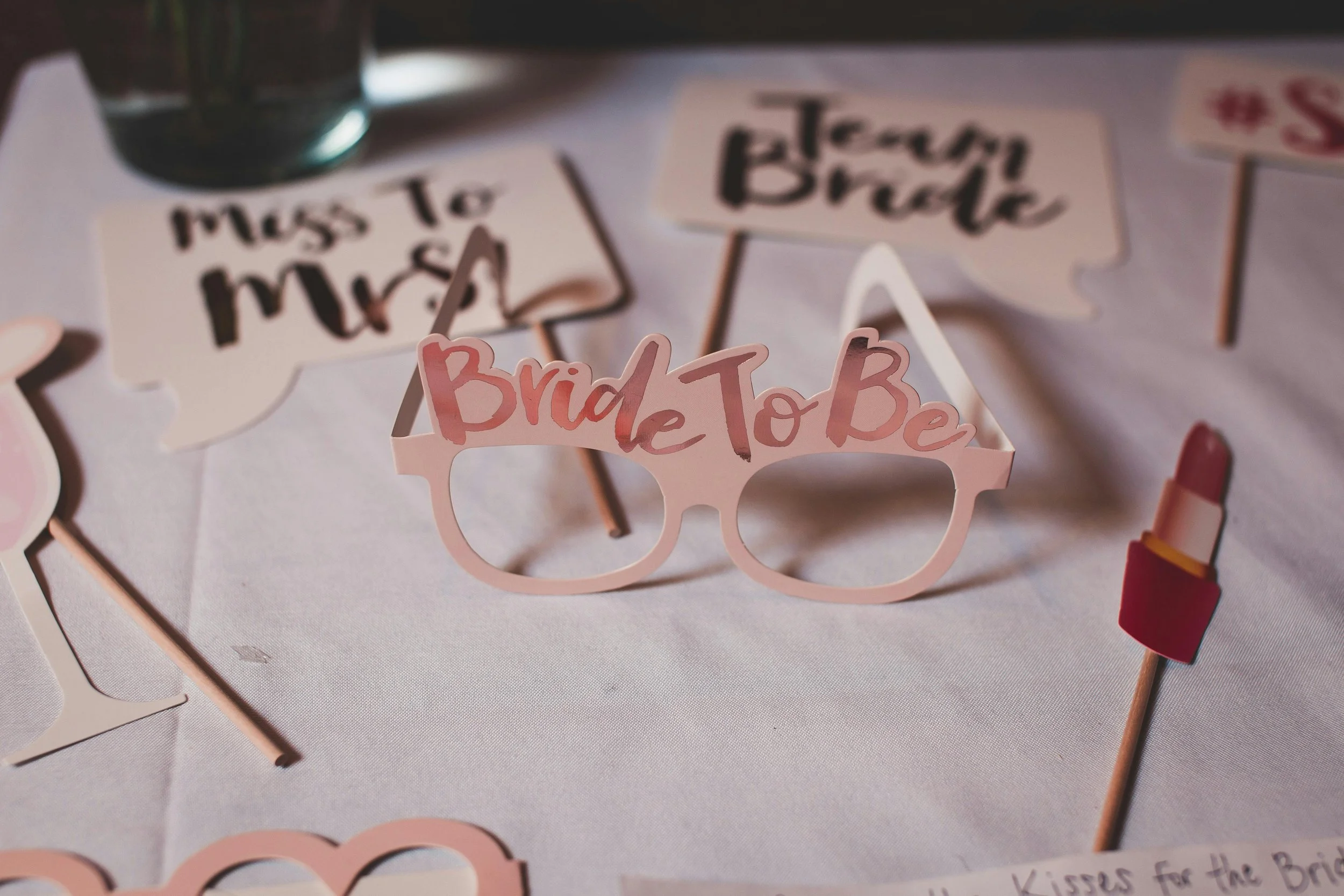 Decorative pink paper glasses with the words 'Bride To Be' cut out on the lenses, surrounded by wedding-themed signs and props on a white table.