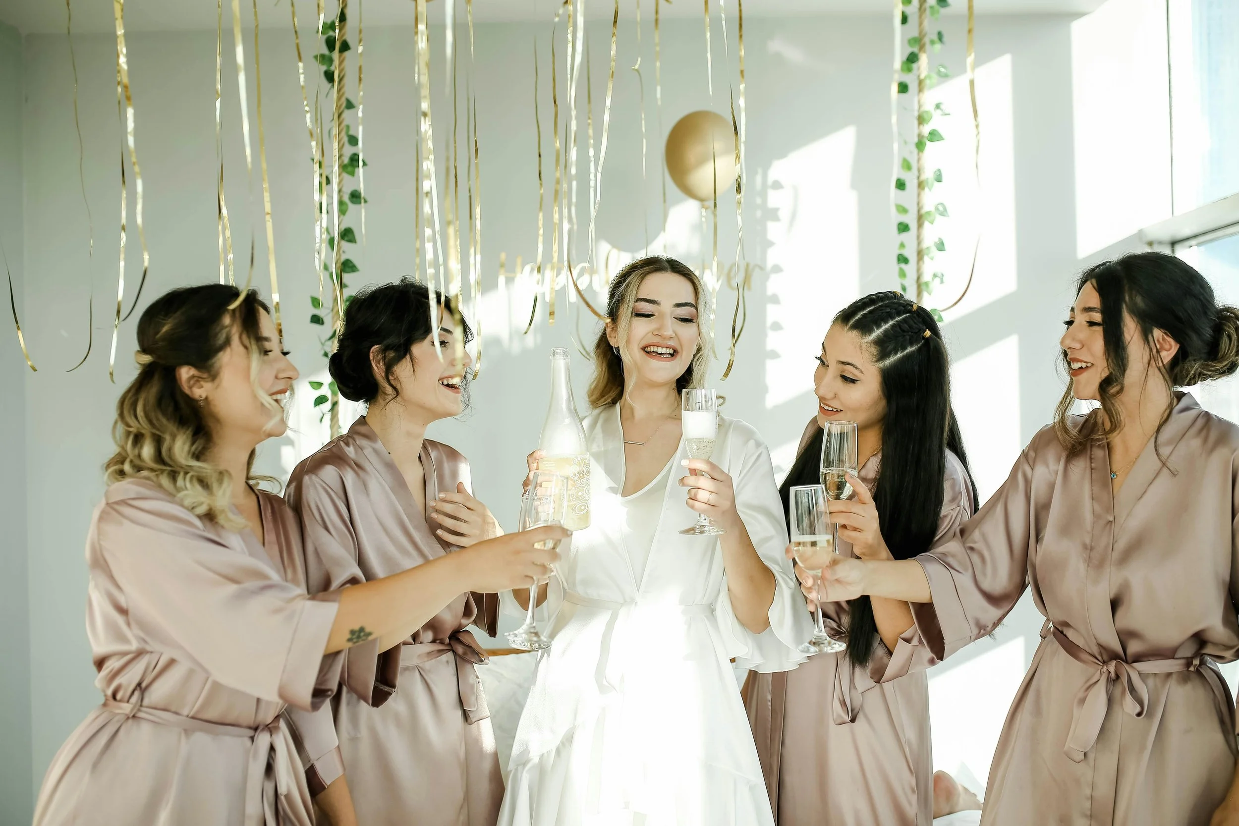Group of women celebrating with drinks in a decorated room, some wearing matching satin robes, indicating a bridal or bridal shower event.
