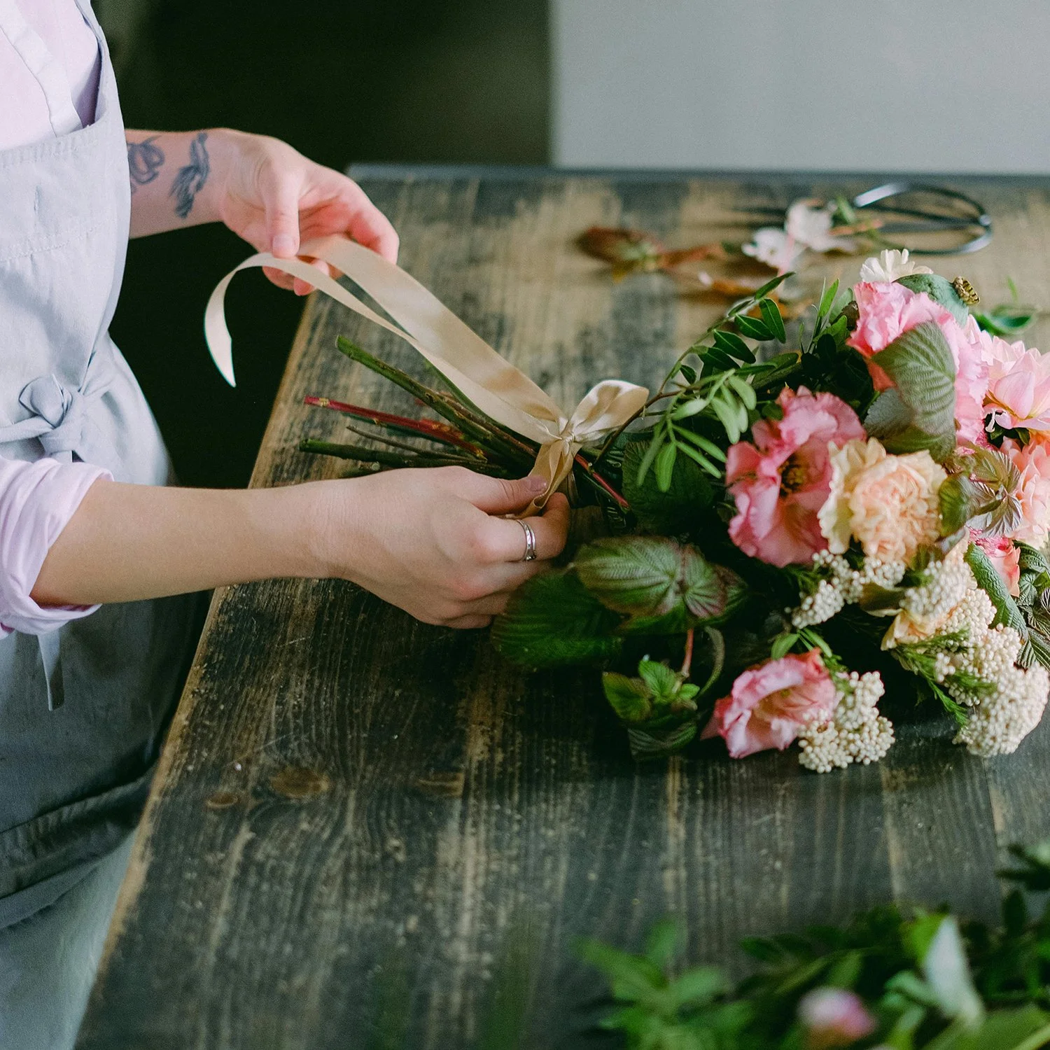 Person tying a beige ribbon on a bouquet of pink and white flowers on a rustic wooden table.