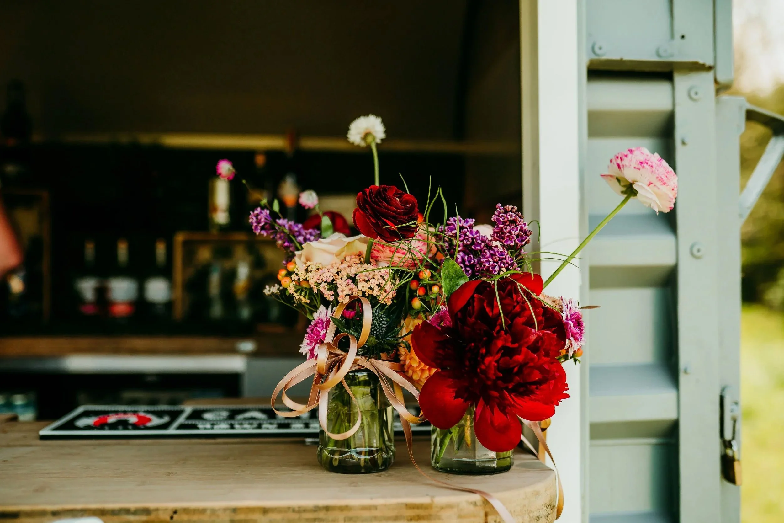 A bouquet of colorful flowers in a glass jar on a wooden countertop with a dark background and some kitchen items.