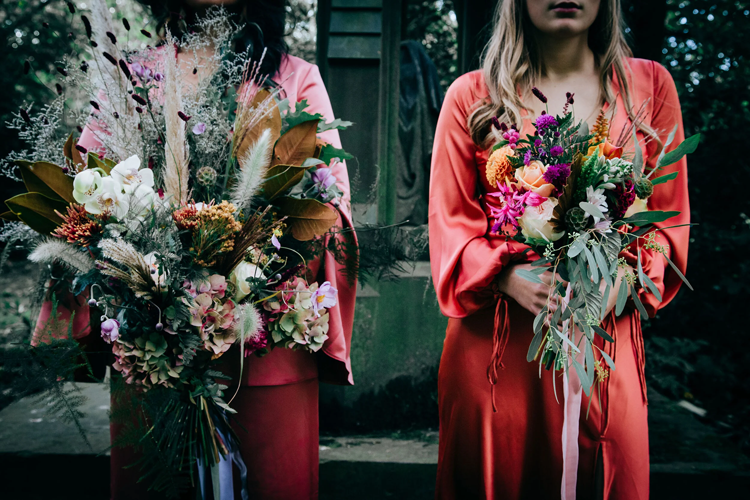 Two women wearing coral-colored dresses holding large bouquets of mixed flowers, including roses, orchids, and greenery, outdoors with a green wooden structure and dark trees in the background.