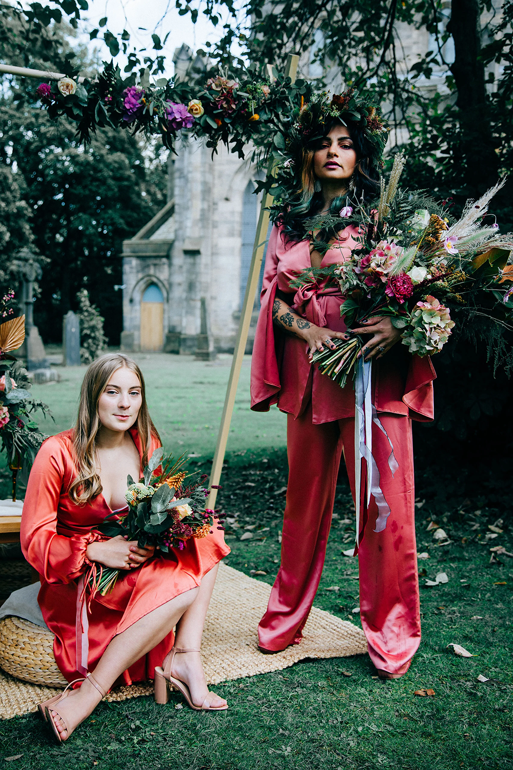 Two women in vibrant pink dresses pose outdoors with floral bouquets, one standing with a large bouquet and a floral crown, the other sitting on a woven stool holding her bouquet, with a historic stone building in the background.