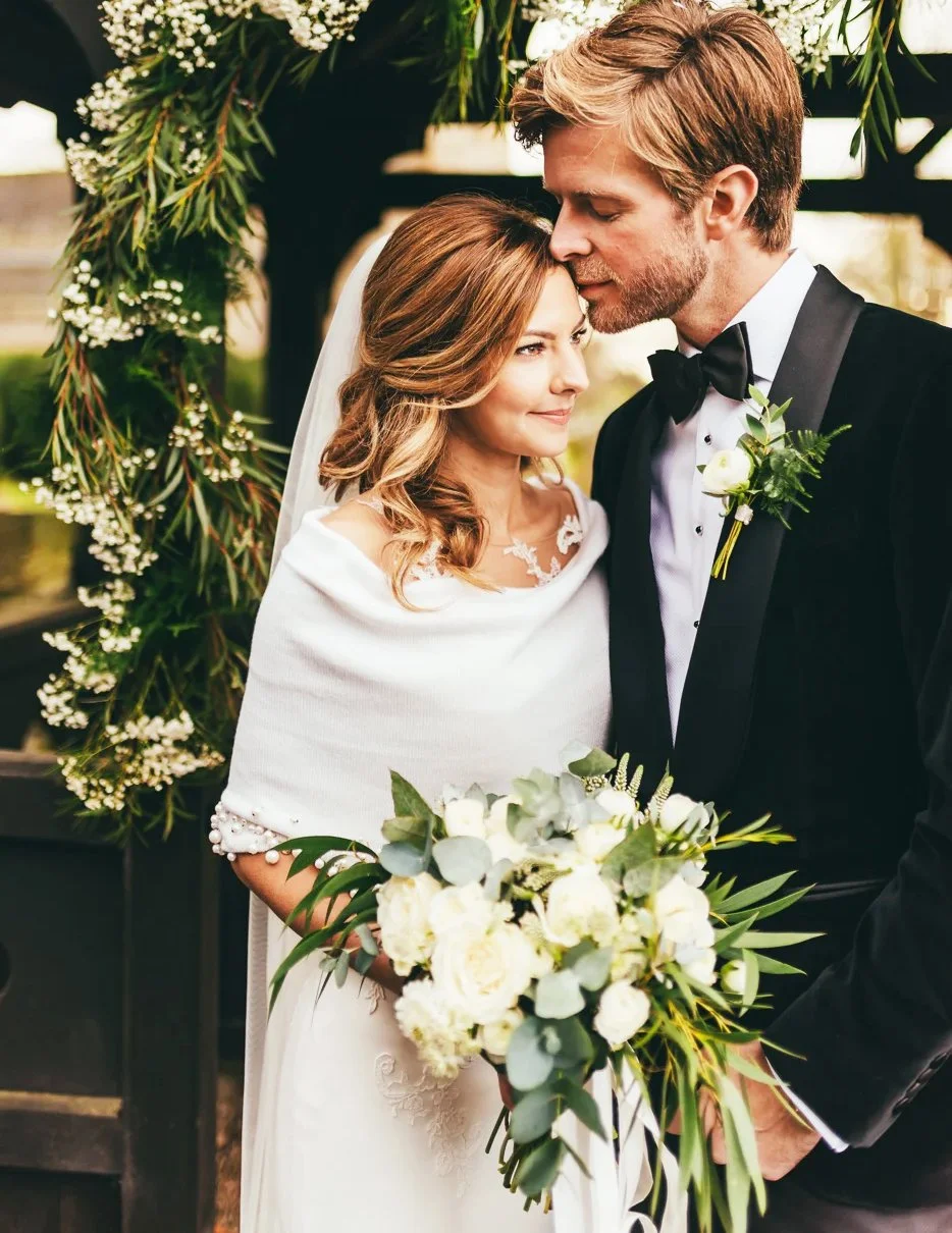 A bride and groom at their wedding, standing close under a floral arch. The bride has shoulder-length, wavy brown hair and is wearing a white dress with lace details and a veil. The groom has light brown hair, a beard, and is dressed in a black tuxedo with a bow tie. They are holding a bouquet of white flowers and greenery.