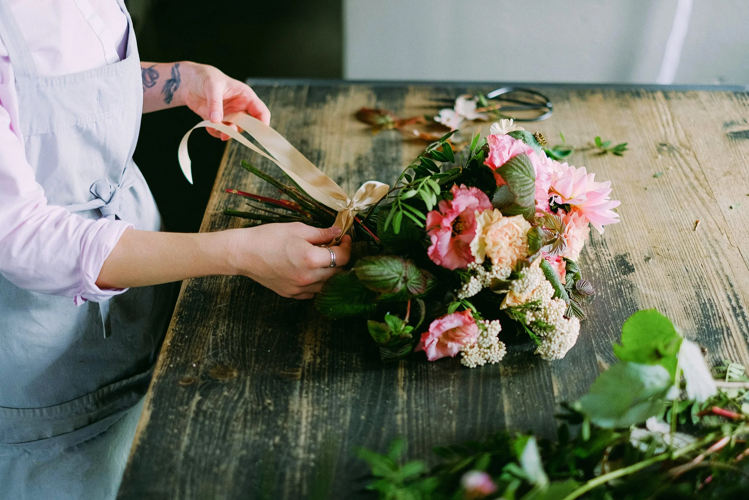 A person arranging a bouquet of pink and cream flowers with green leaves on a wooden table, with floral scissors and stems nearby.