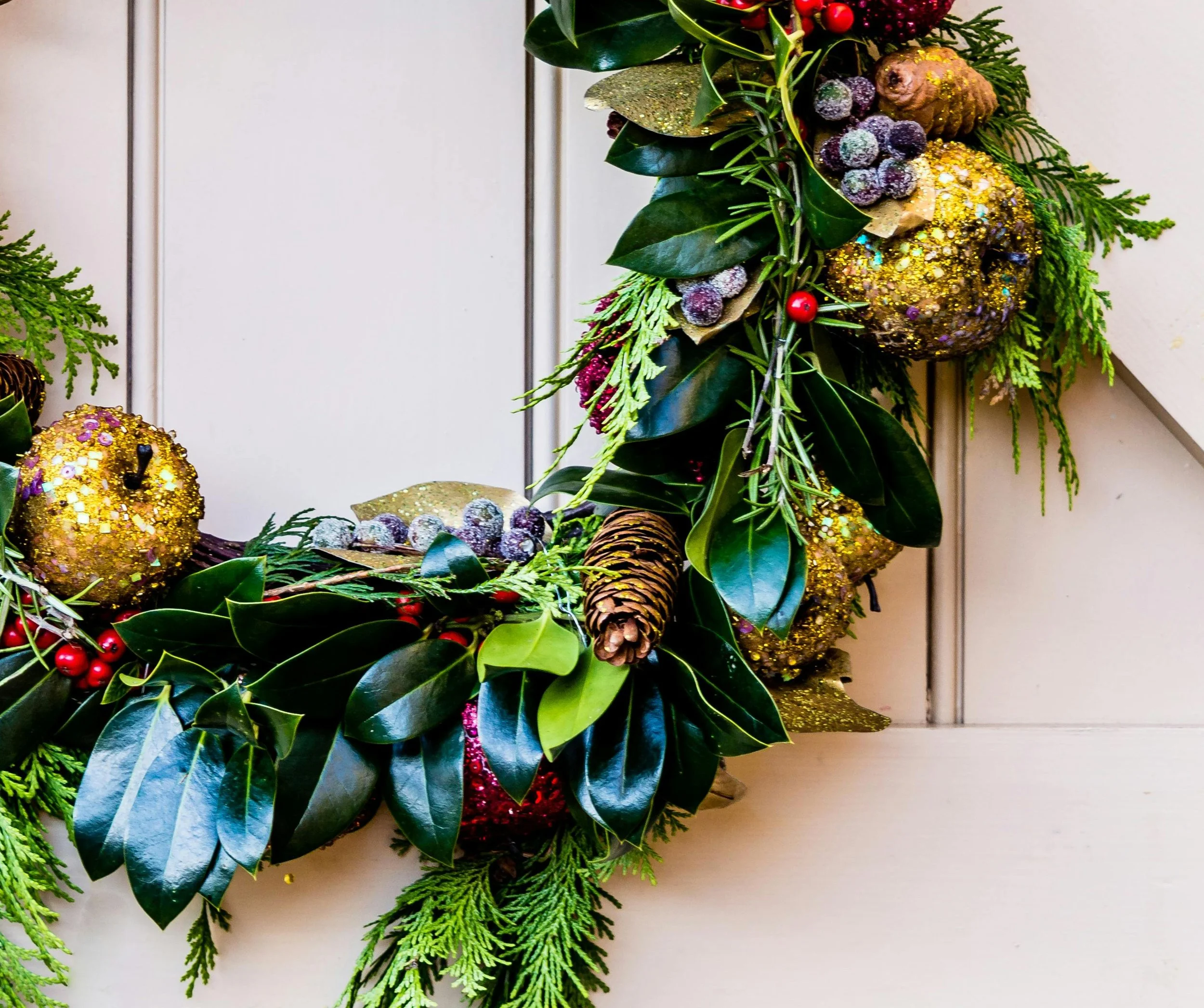 Close-up of a Christmas wreath with green leaves, small red berries, pinecones, and decorated gold and purple ornaments hanging on a white wall.