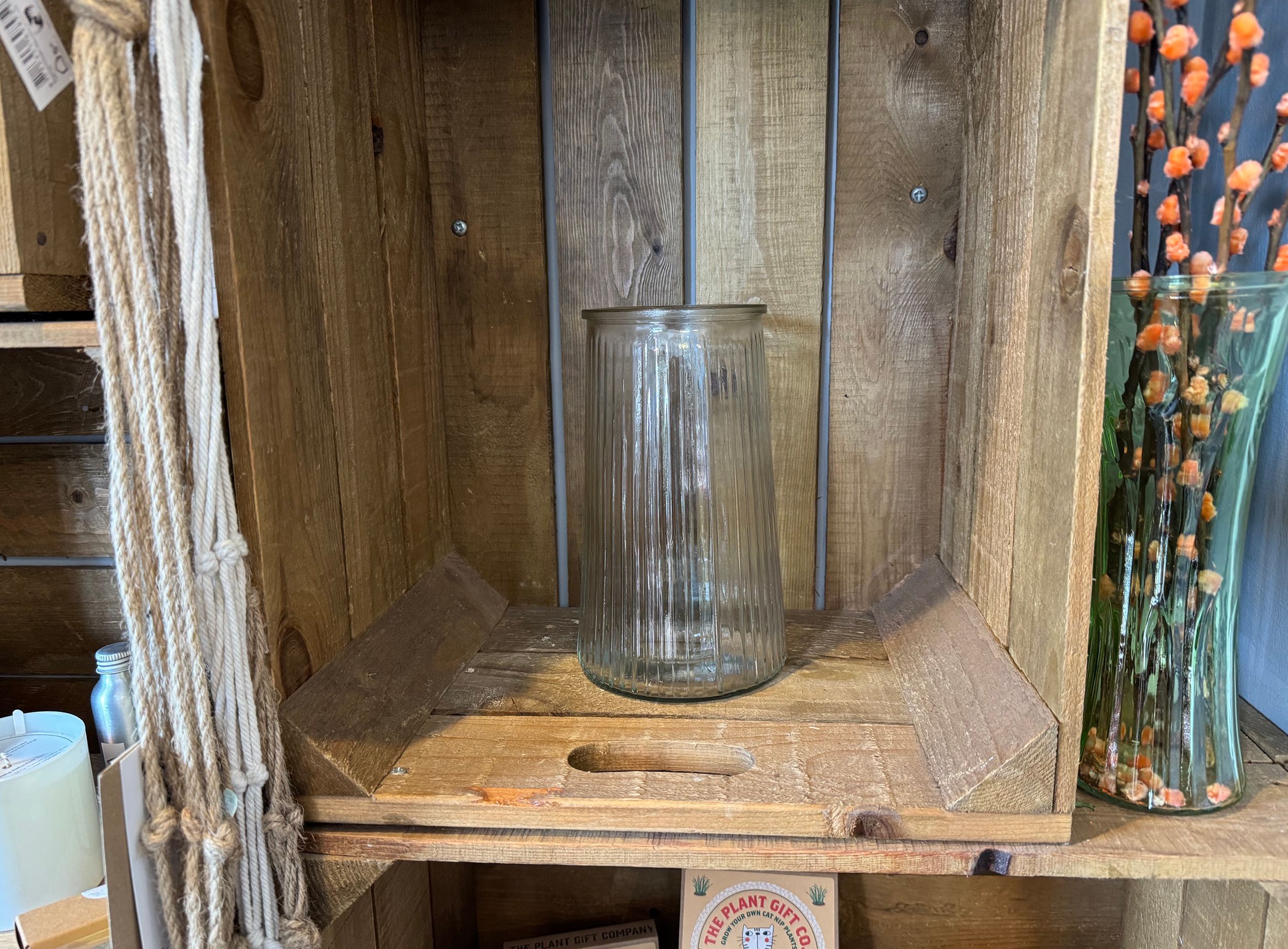A wooden display shelf with a tall, closed, clear glass vase in the center, flanked by what appears to be dried orange and yellow flower arrangements in glass containers on the right side.