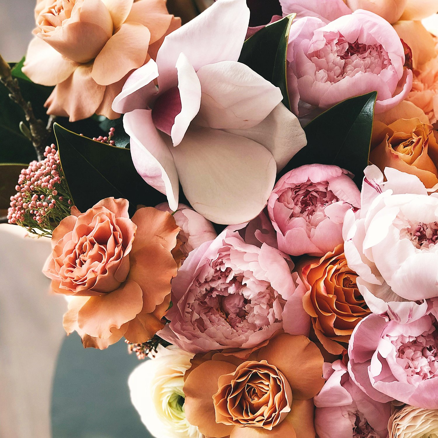 A close-up of a bouquet containing blush pink, peach, and cream roses, pink peonies, and greenery.