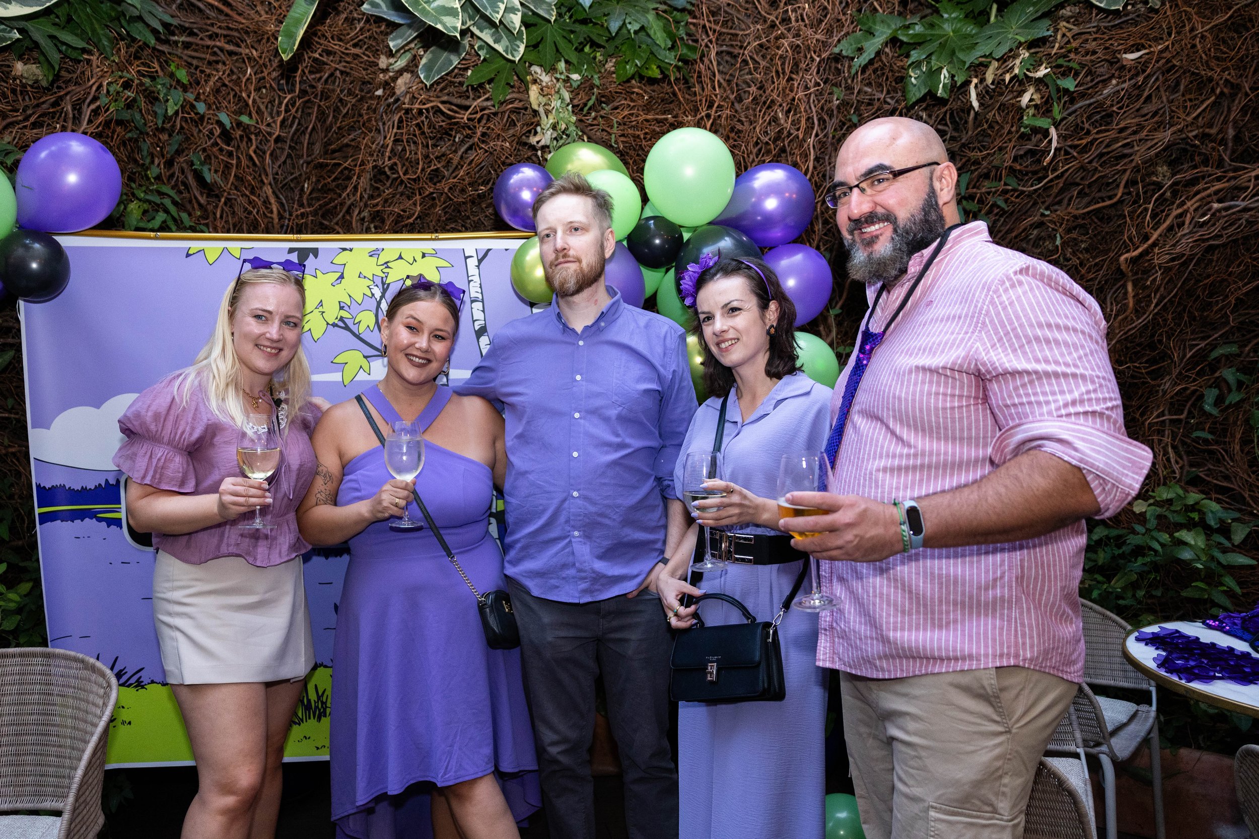 Group of five people at a party, standing in front of a decorated backdrop with purple, green, black balloons, and a cartoonish nature scene.