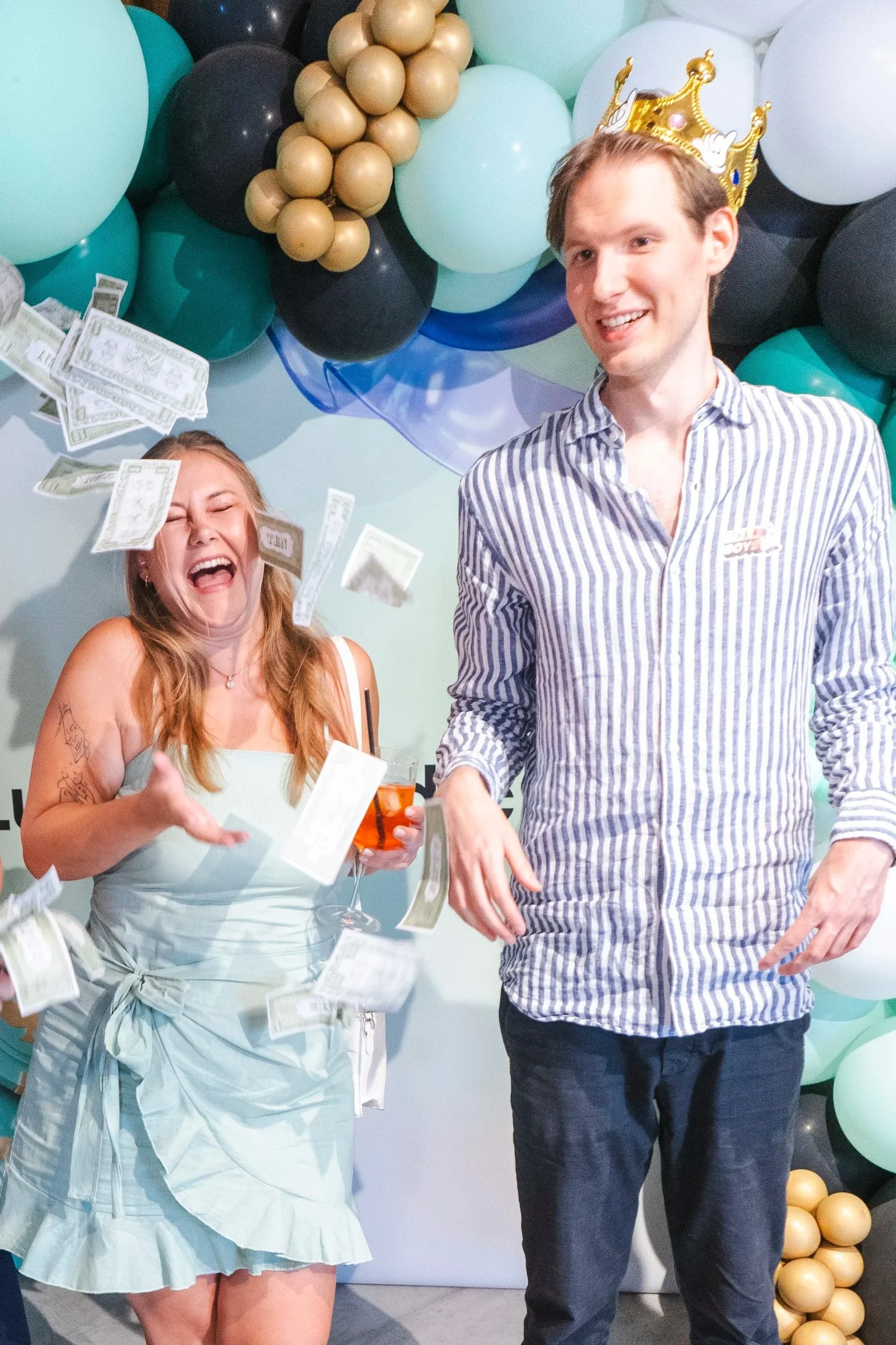 Three people celebrating at a party with balloons and confetti, smiling and throwing money in the air.