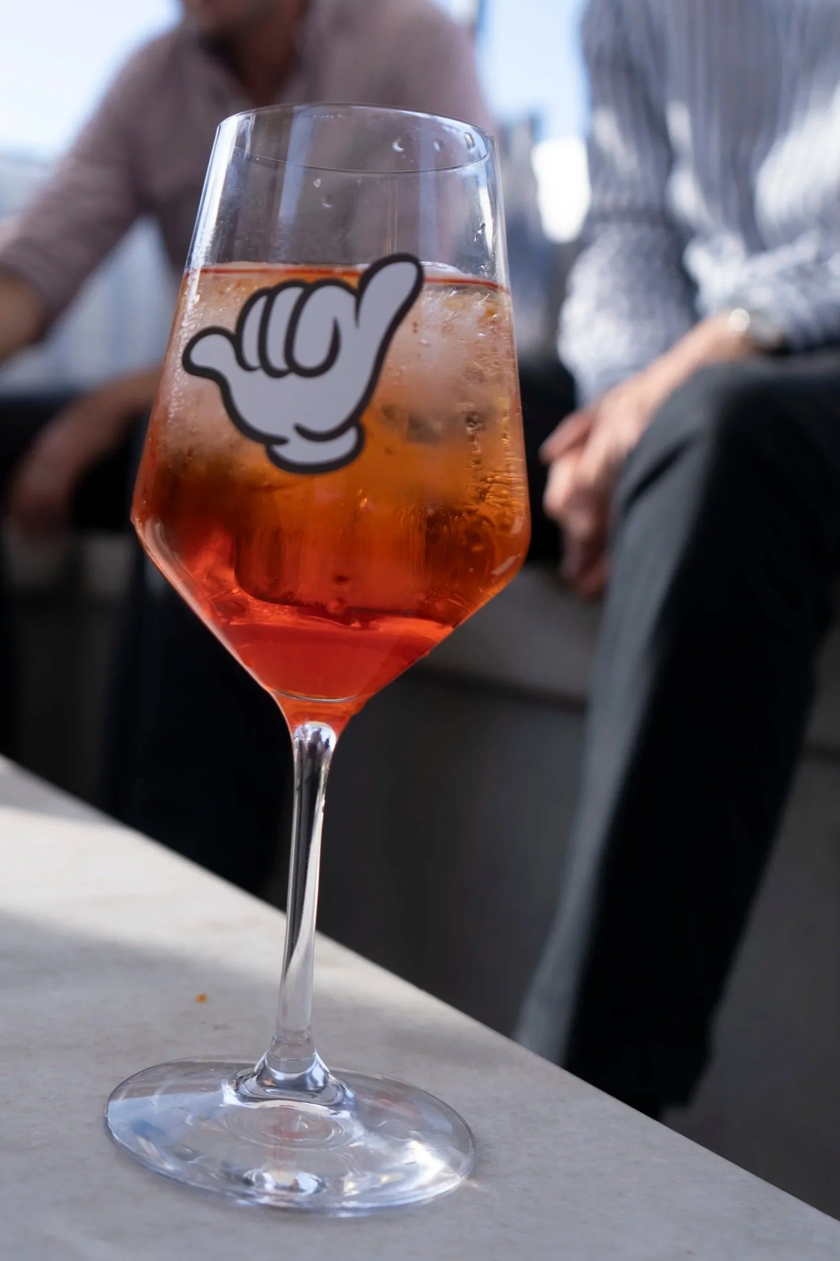 A glass of Aperol Sprtiz with a sticker of a cartoon hand making a shaka sign, on a table near seated people in the background during daytime.