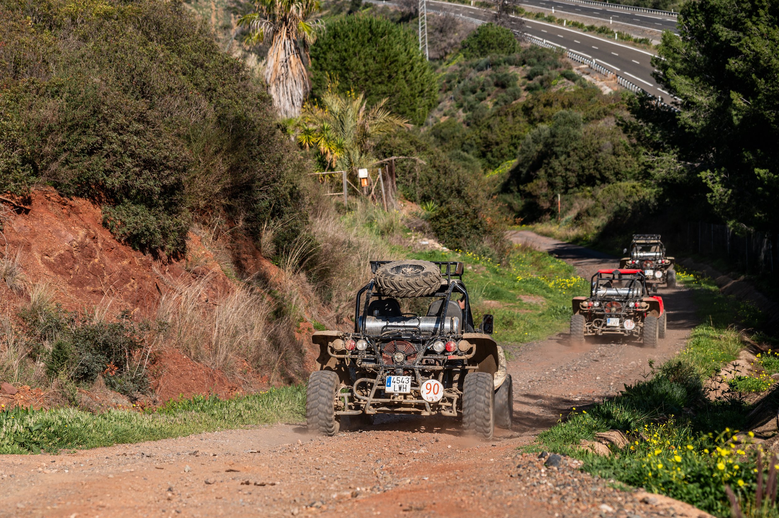 Three off-road vehicles driving on a dirt trail through a hilly, green landscape with trees and shrubs, under a clear sky.