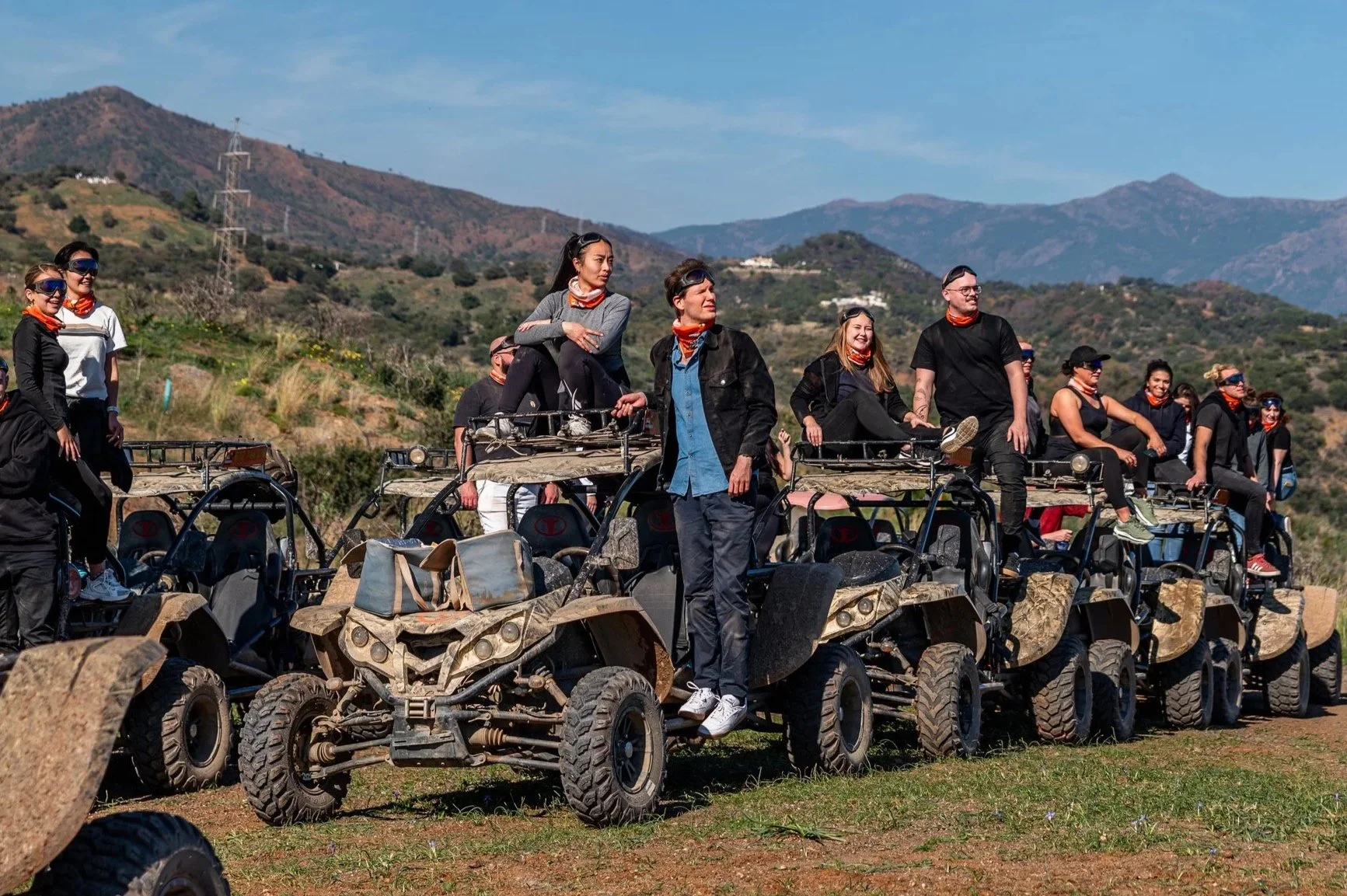 A group of people riding all-terrain vehicles in a mountainous outdoor setting, some sitting on the vehicles and others standing or sitting on top, with mountains and clear sky in the background.