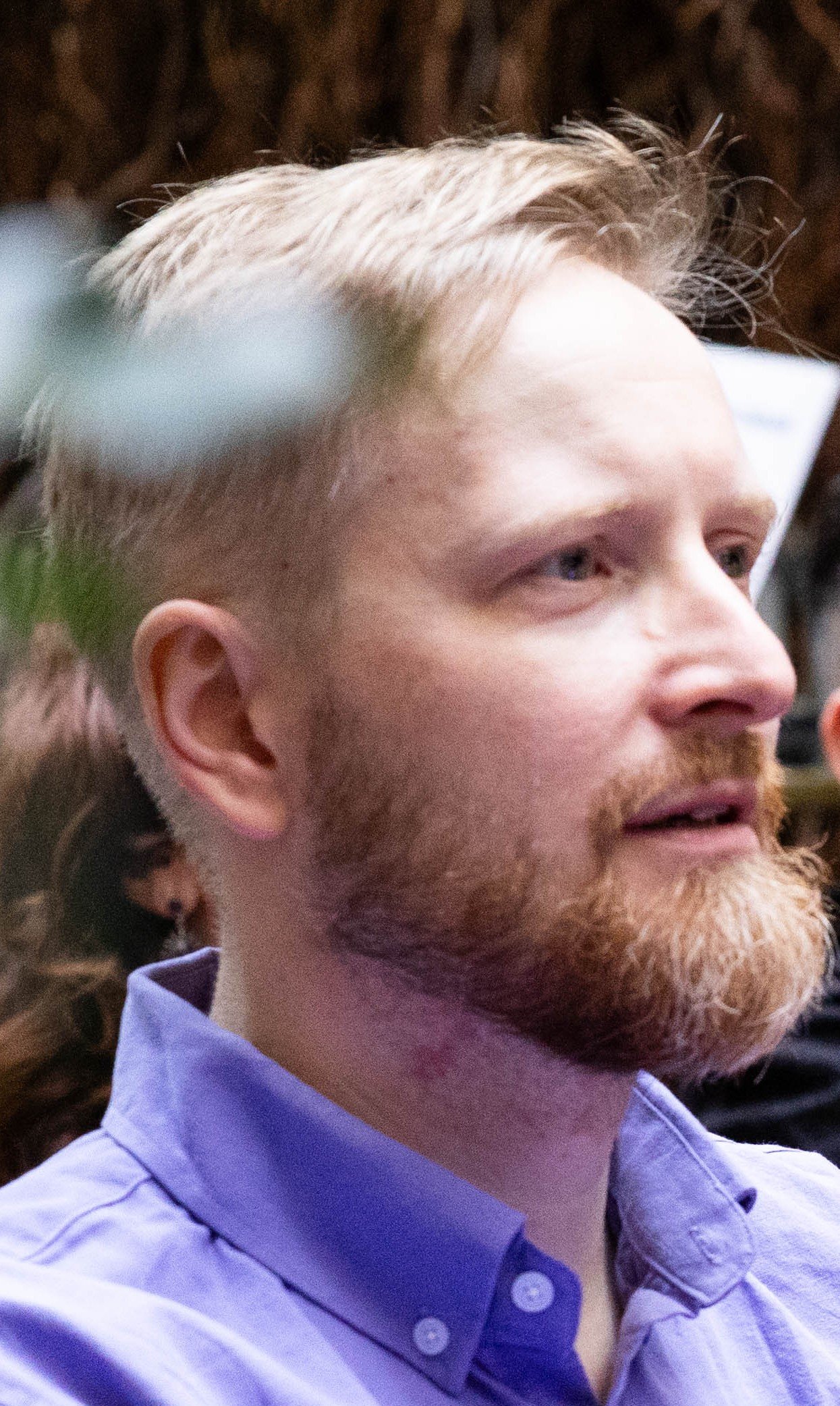 A side profile of a man with light-colored hair and a beard, wearing a light purple collared shirt.