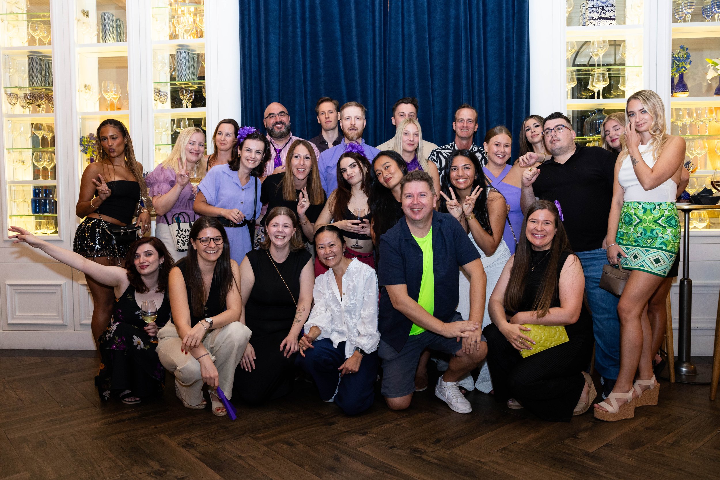 A large group of people at a celebration or party in an elegant room with glassware and decorative items on display, posing for a group photo, smiling and making peace signs.