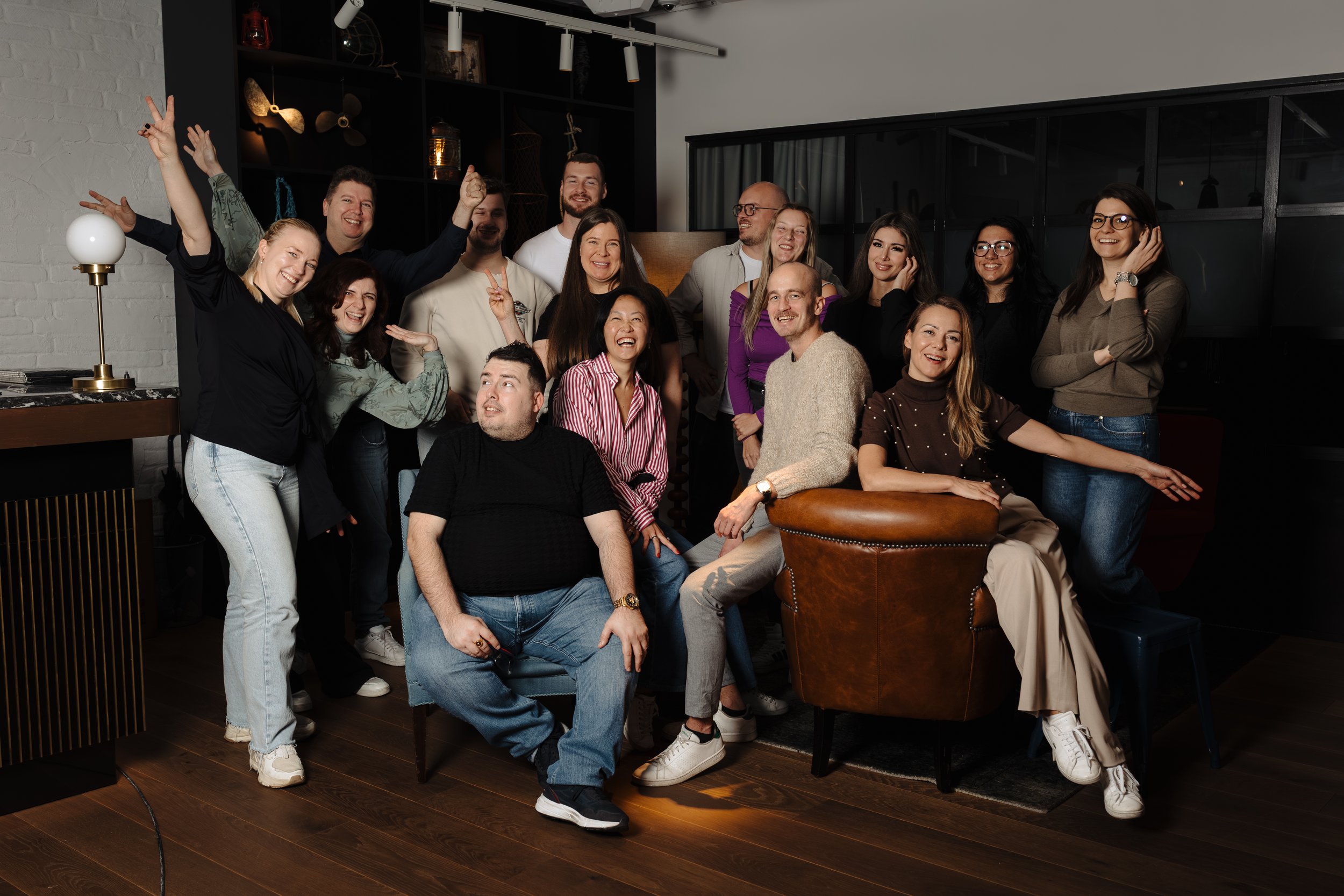 A group of 15 diverse people gathered in a modern living room, smiling and posing for a group photo. Some are standing, others seated, with a mix of casual and cheerful expressions.