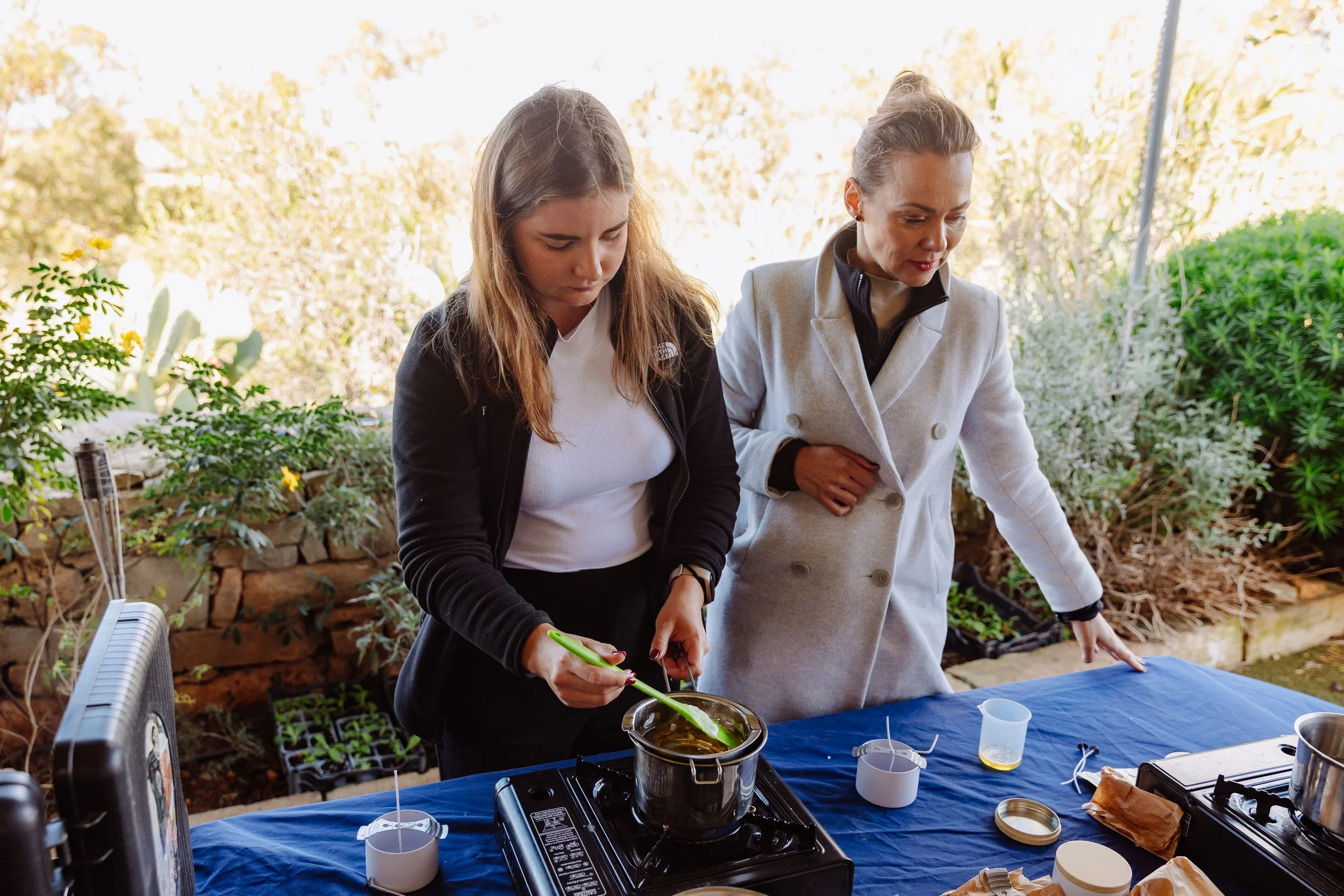 Two women preparing food outdoors at a table covered with a blue cloth, with plants and rocks in the background.