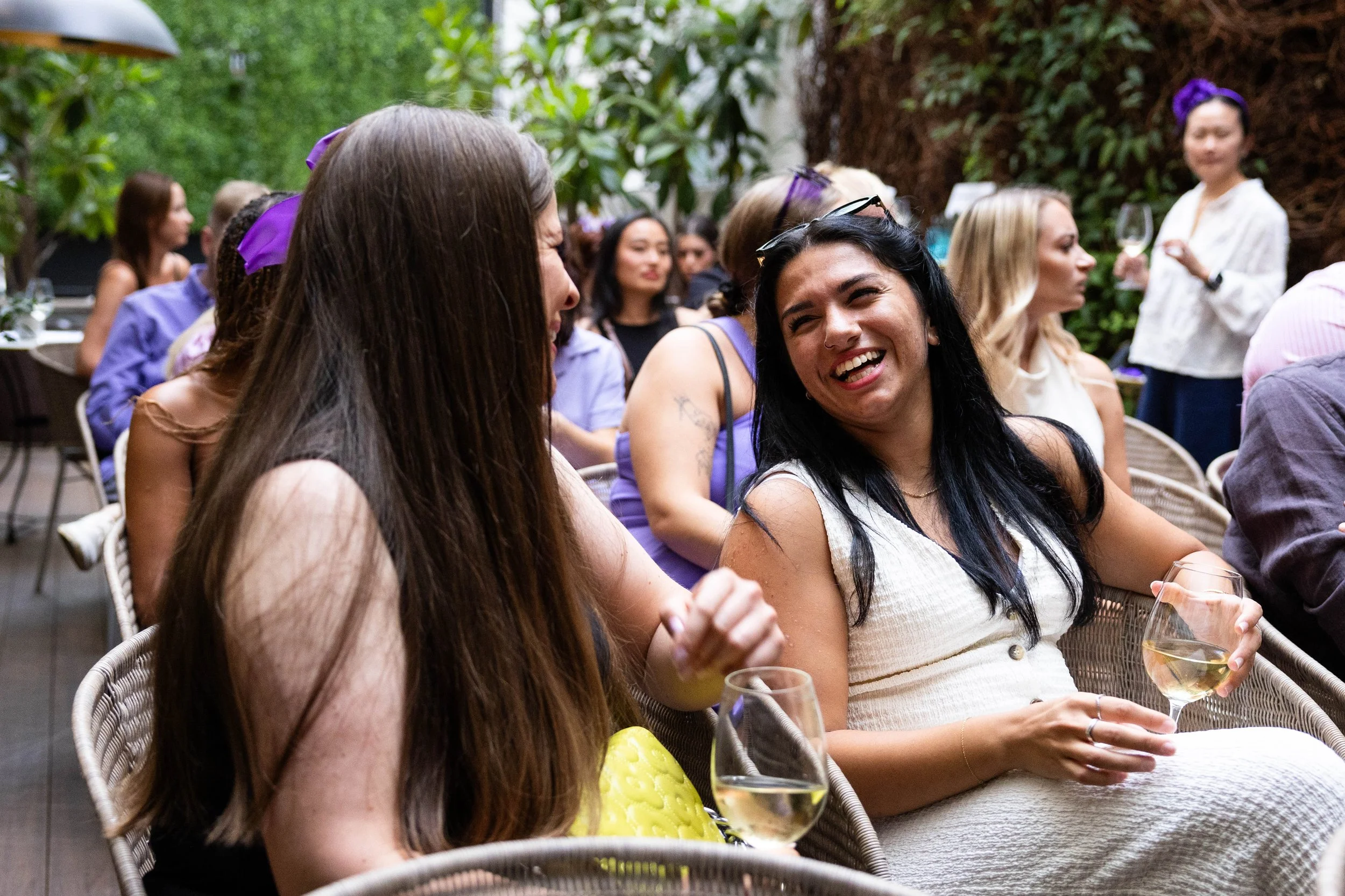 Group of women at an outdoor social gathering, some holding wine glasses, smiling, and talking, with lush greenery in the background.