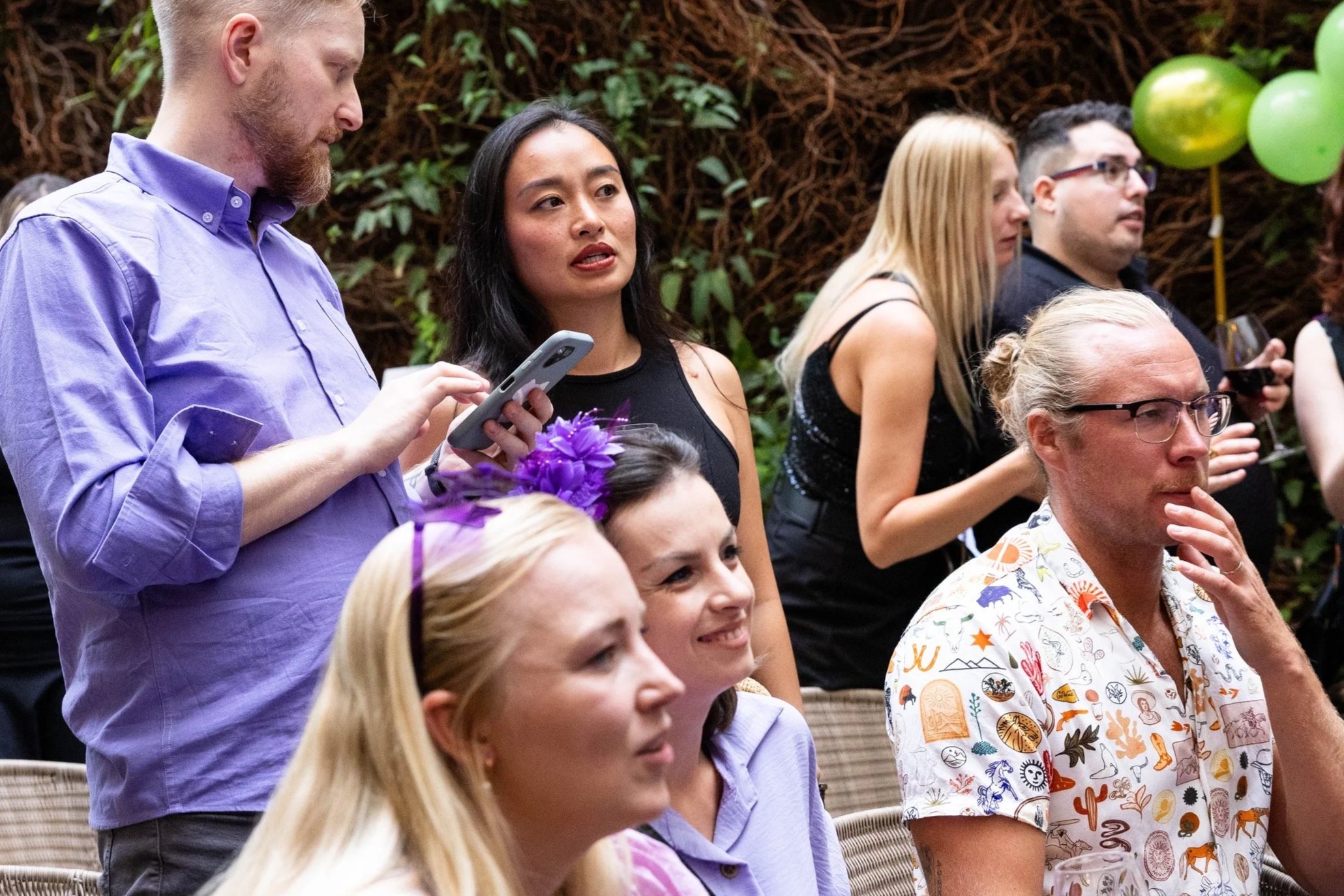 People attending an outdoor event with green balloons, some seated and others standing, engaged in conversation or observing.